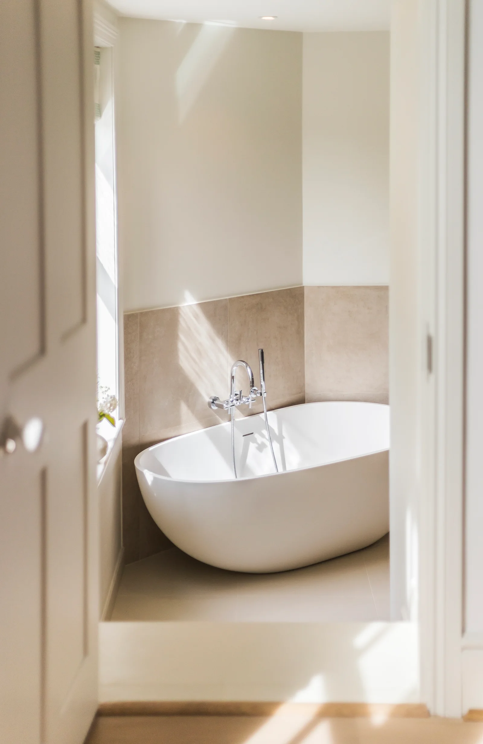 A white oval freestanding bathtub sits in a tiled bathroom corner next to a window with natural light.