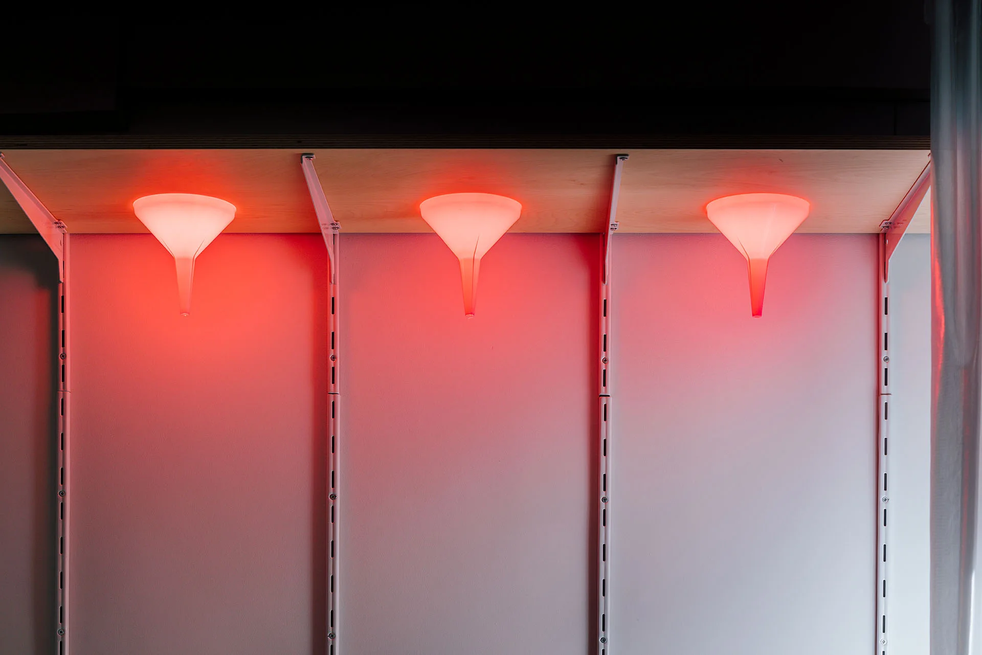 Three funnel-shaped lights cast a red glow underneath a light wood shelf supported by white adjustable wall tracks.