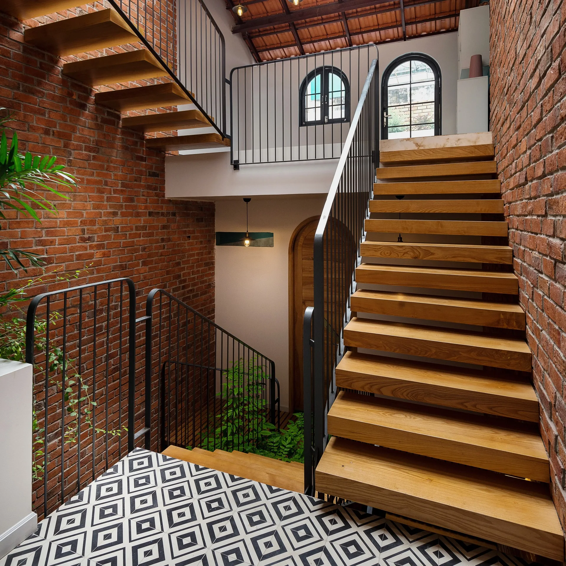 An interior multi-level staircase with floating wooden treads, black metal railings, exposed red brick walls, and geometric tiled flooring.
