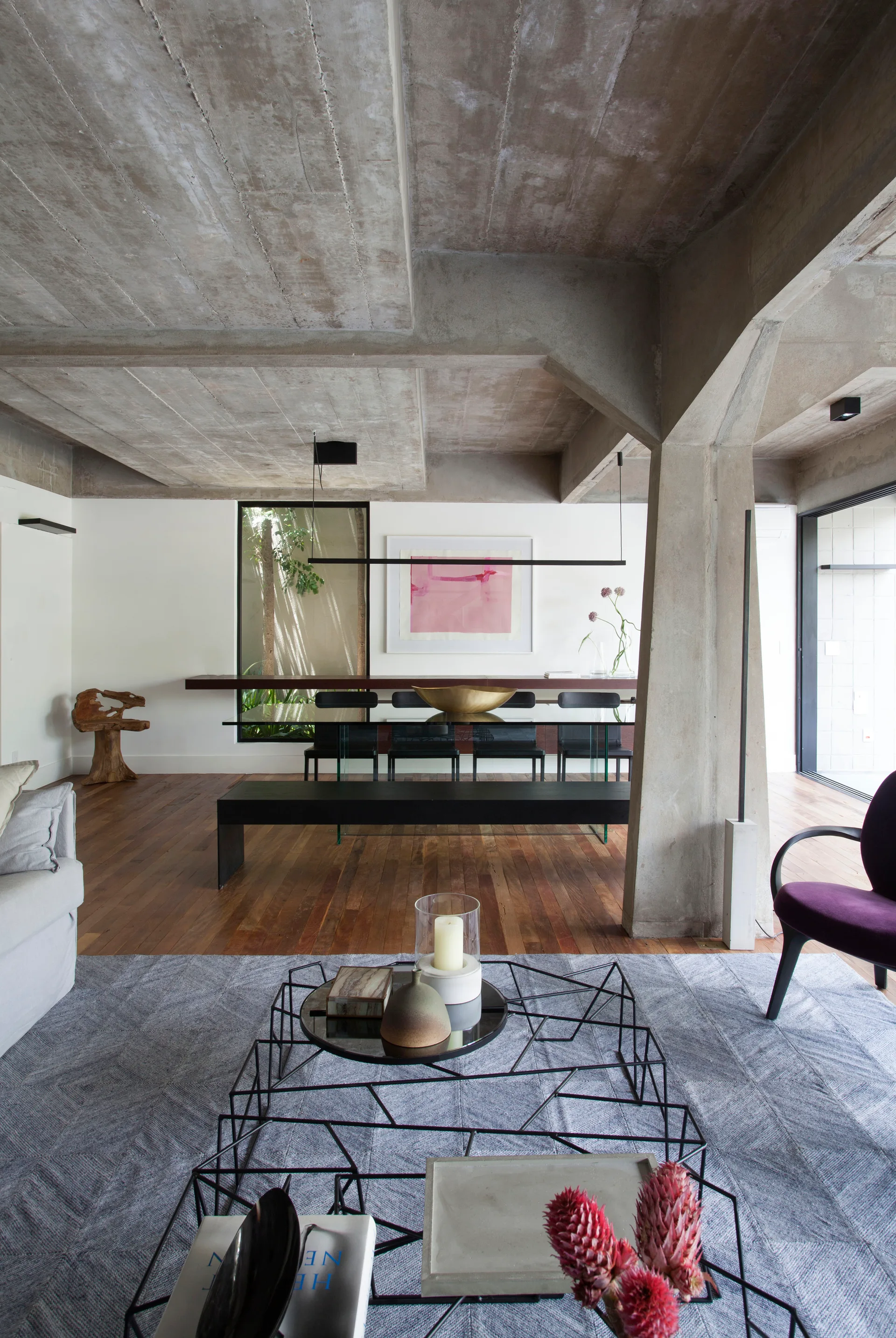 An open-plan living and dining area featuring raw concrete ceilings, a glass-top dining table, a black wire coffee table, and wood floors.
