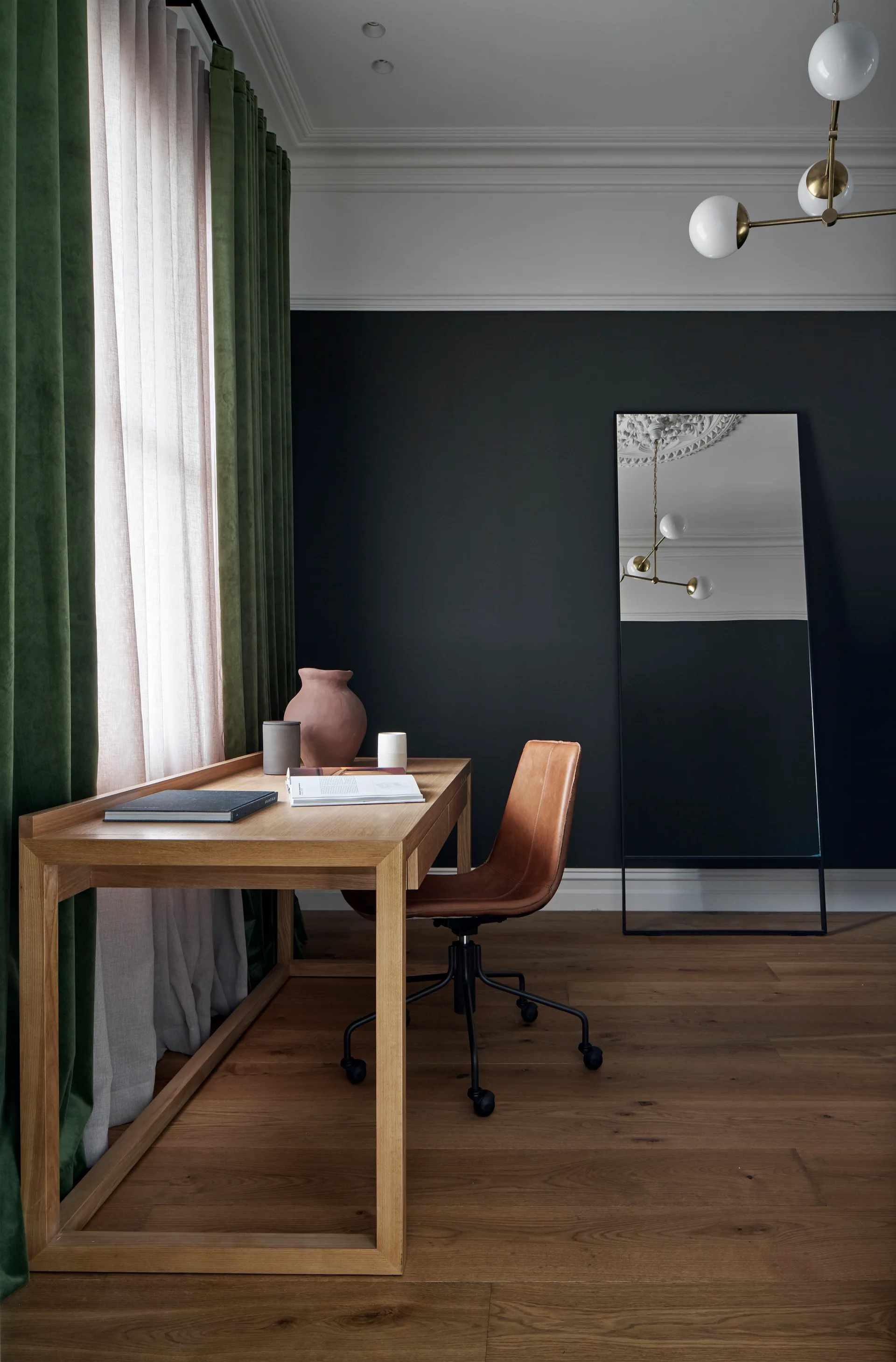 A light wood desk and brown leather chair sit in a home office with dark gray walls, green drapes, and a leaning floor mirror.