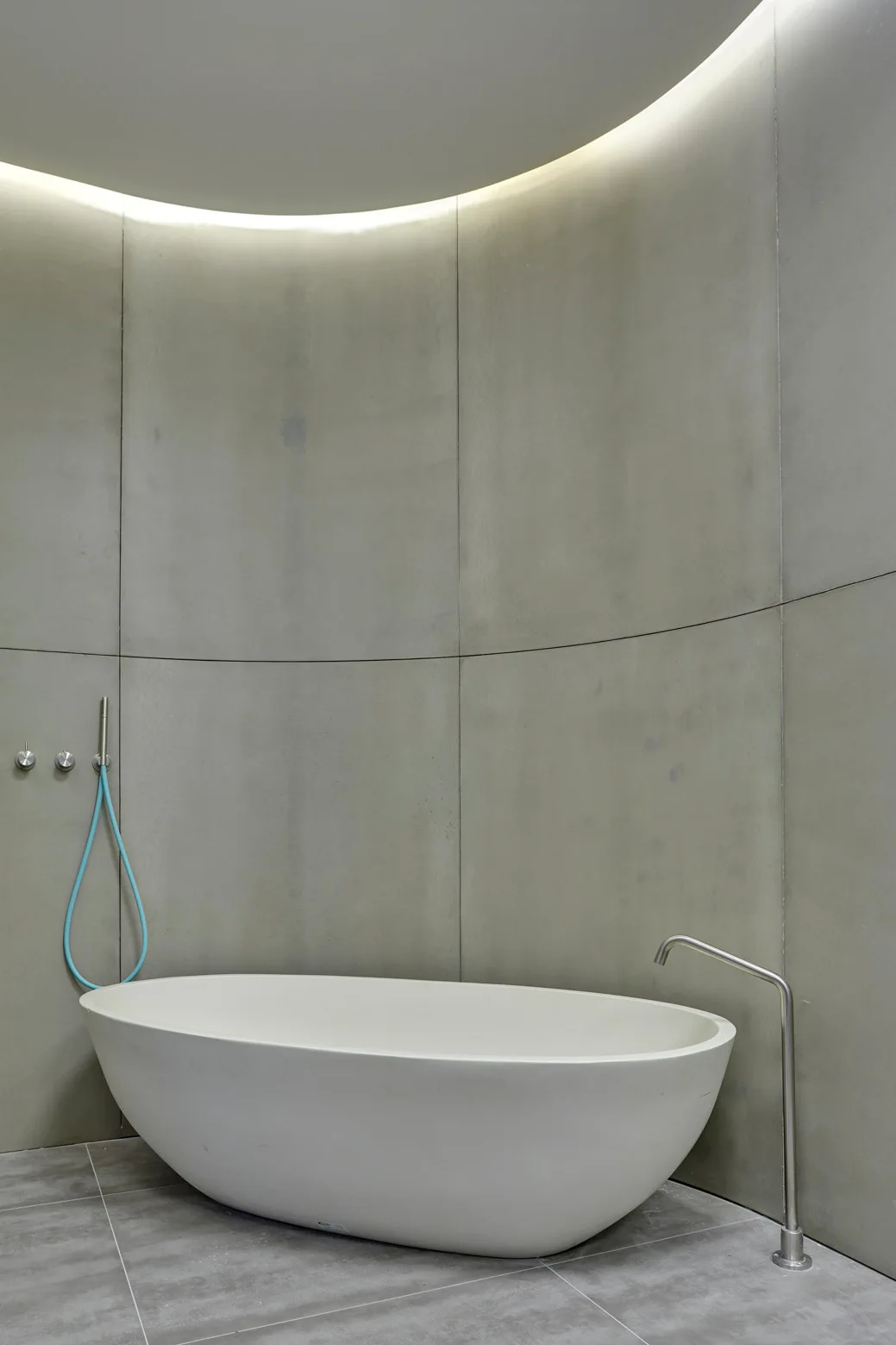 A white freestanding oval bathtub sits against a curved grey concrete wall under soft cove lighting.