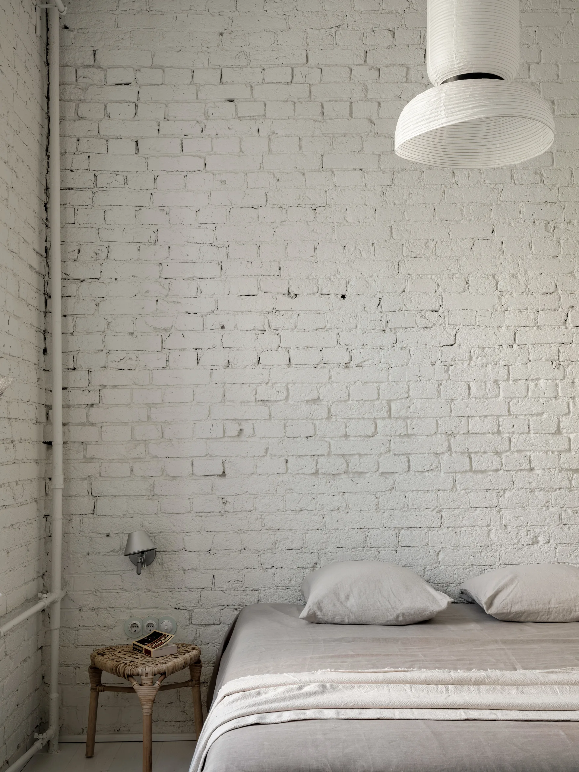 A minimalist bedroom features grey linen bedding against a white painted brick wall next to a small wicker stool and wall sconce.