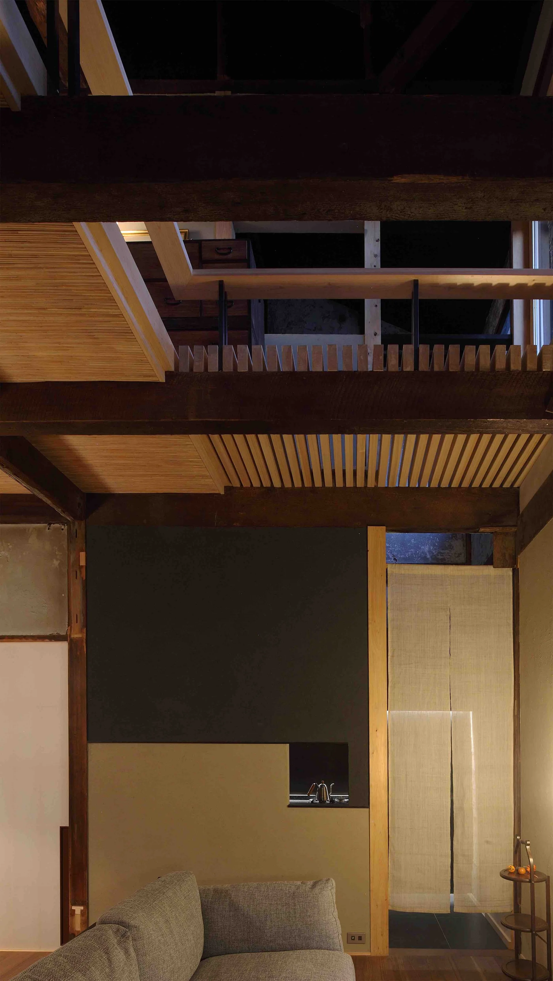 A modern living room features a gray sofa, exposed timber beams, a wood slatted ceiling detail, and a linen noren curtain over a doorway.
