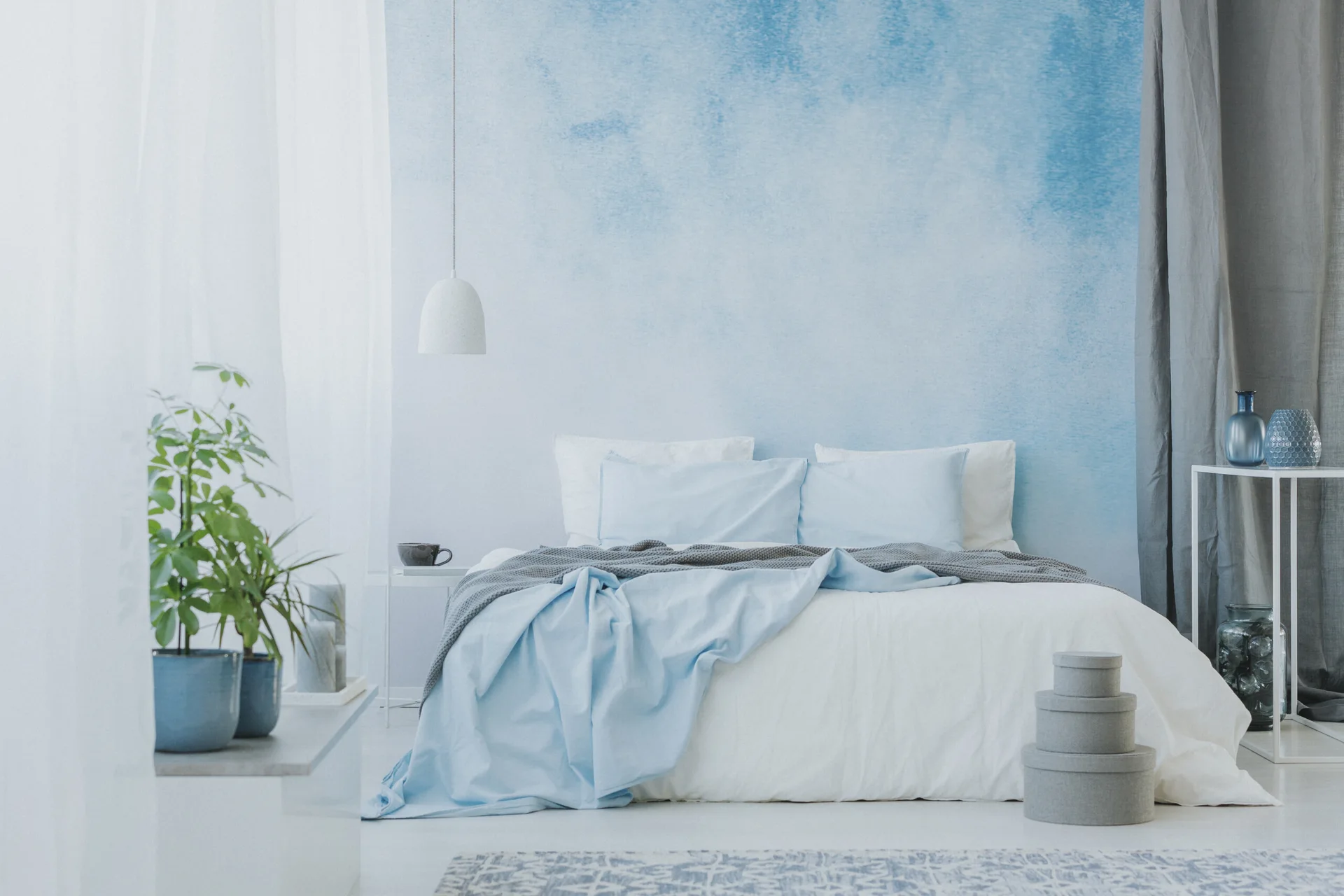 A minimalist bed sits against a blue watercolor ombre wall in a bedroom accented with a white pendant light and a knit throw.