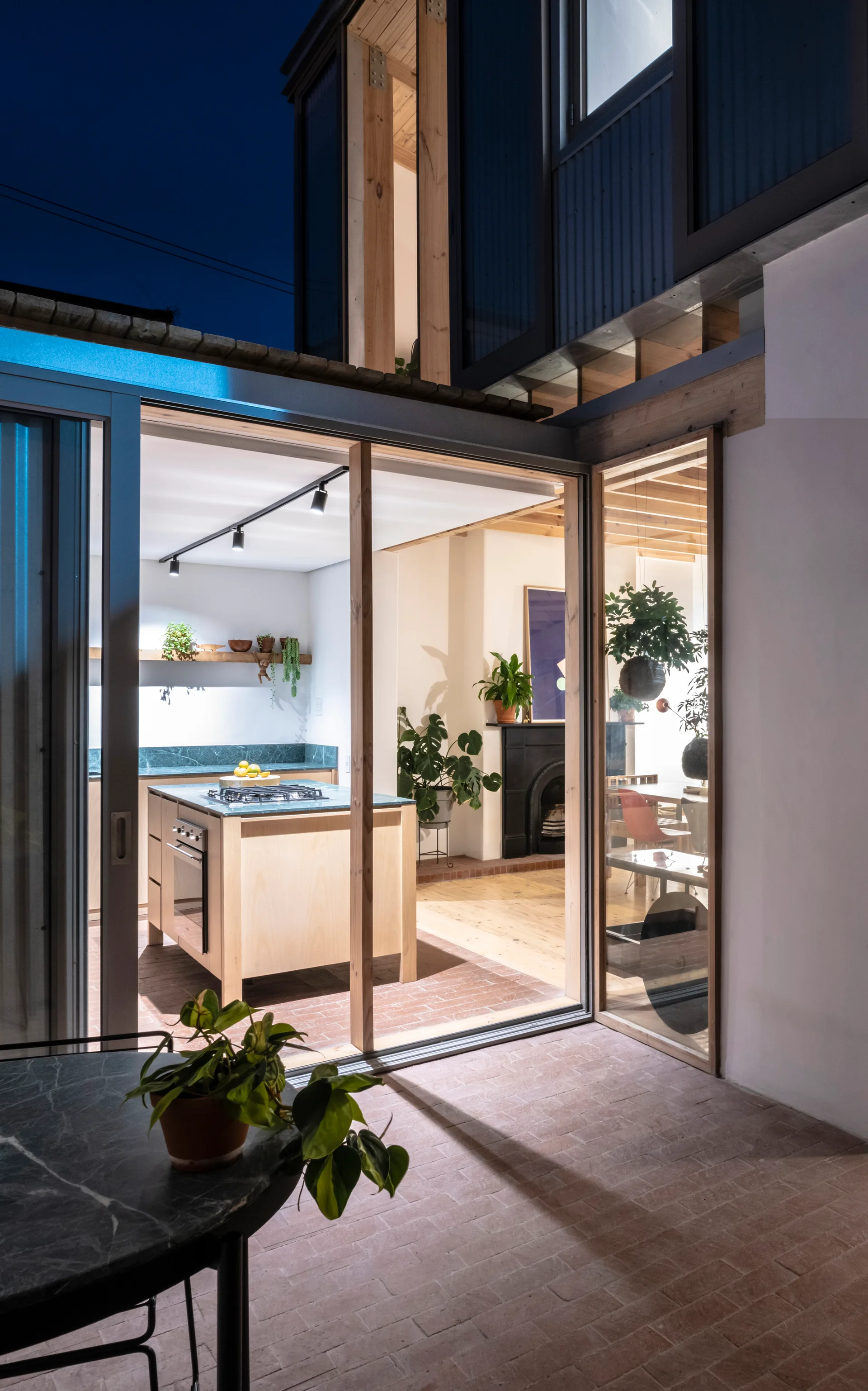 A modern kitchen with light wood cabinets and a stone-topped island opens through large sliding glass doors onto a brick-paved patio.