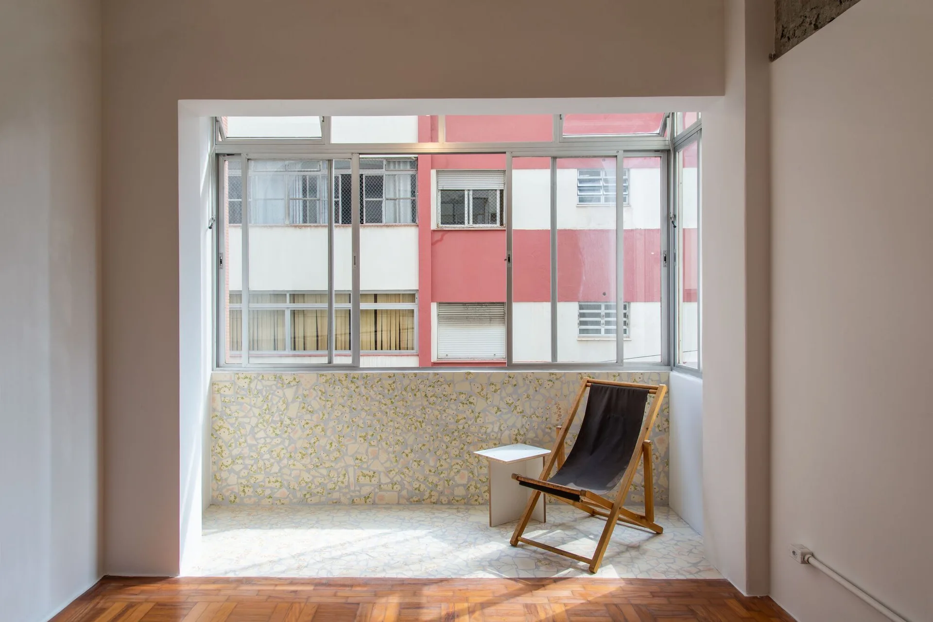 An enclosed balcony features a wooden folding chair on a light terrazzo floor and half-wall next to large metal-framed windows.