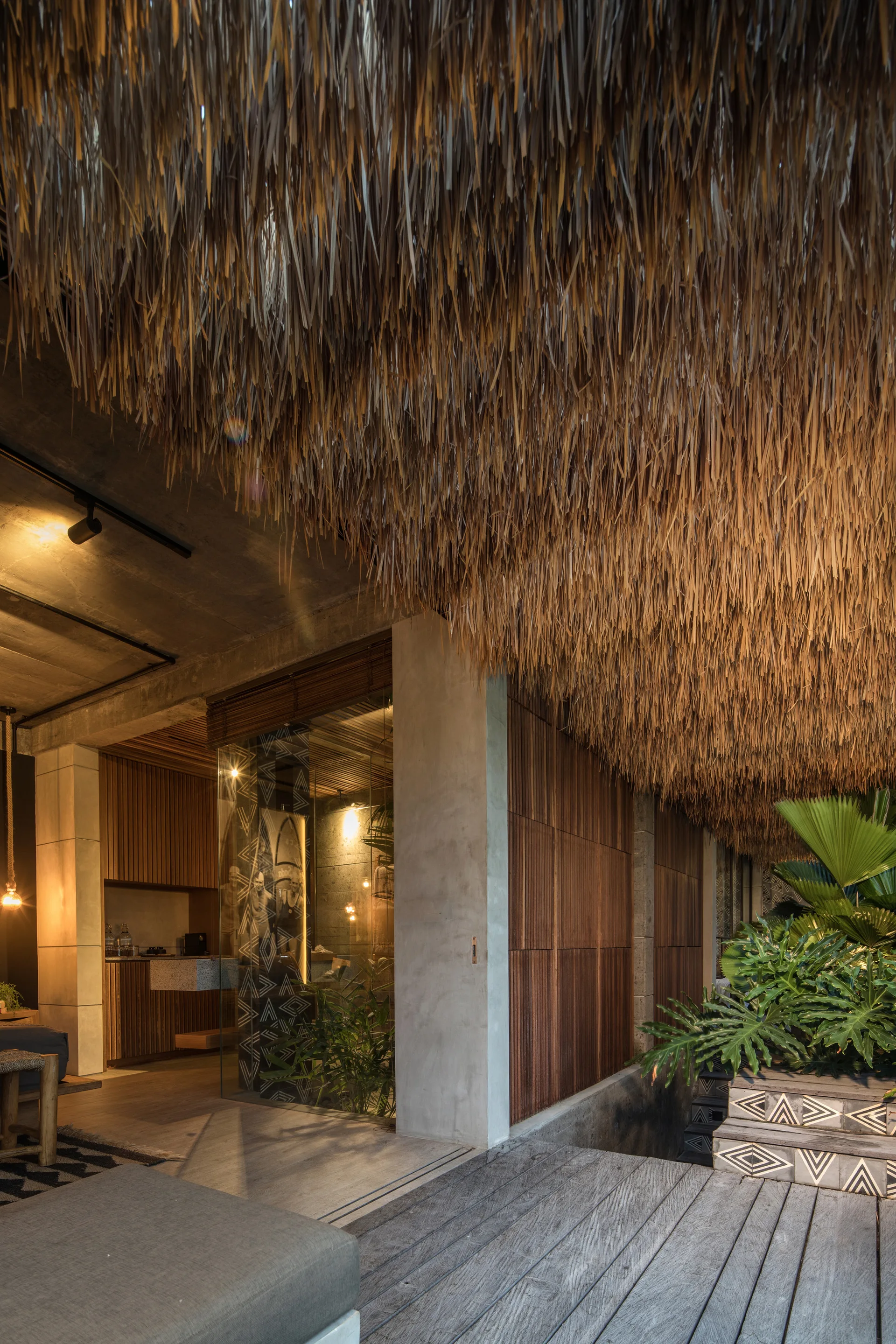 A contemporary tropical patio featuring a thick thatched ceiling, concrete pillars, vertical slatted wood walls, and patterned tile steps.