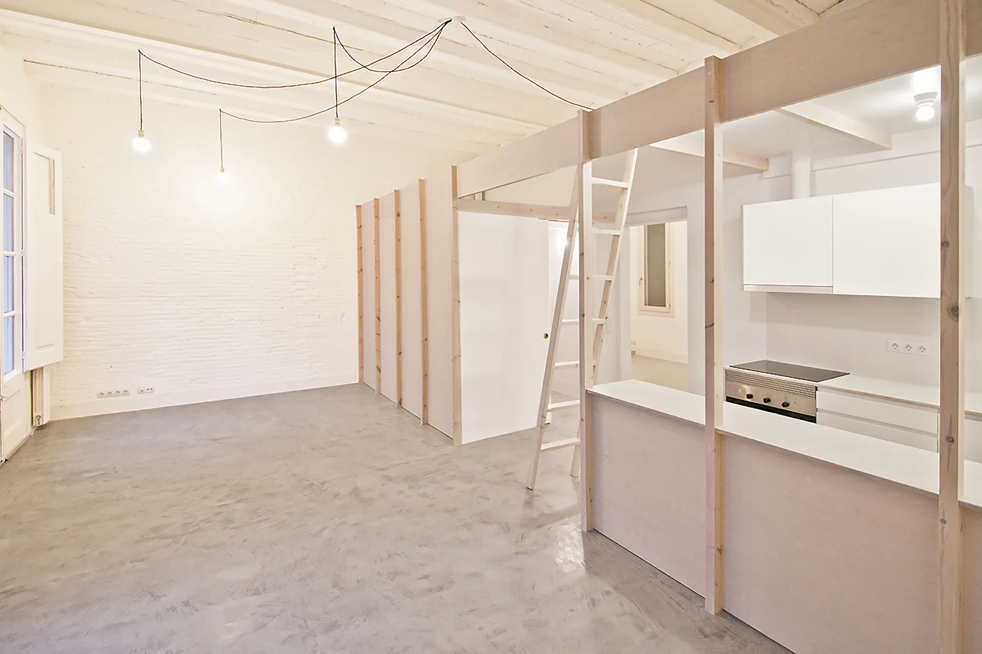 An open-concept studio apartment featuring a grey concrete floor, white painted brick wall, and a wooden loft structure next to a compact kitchen.