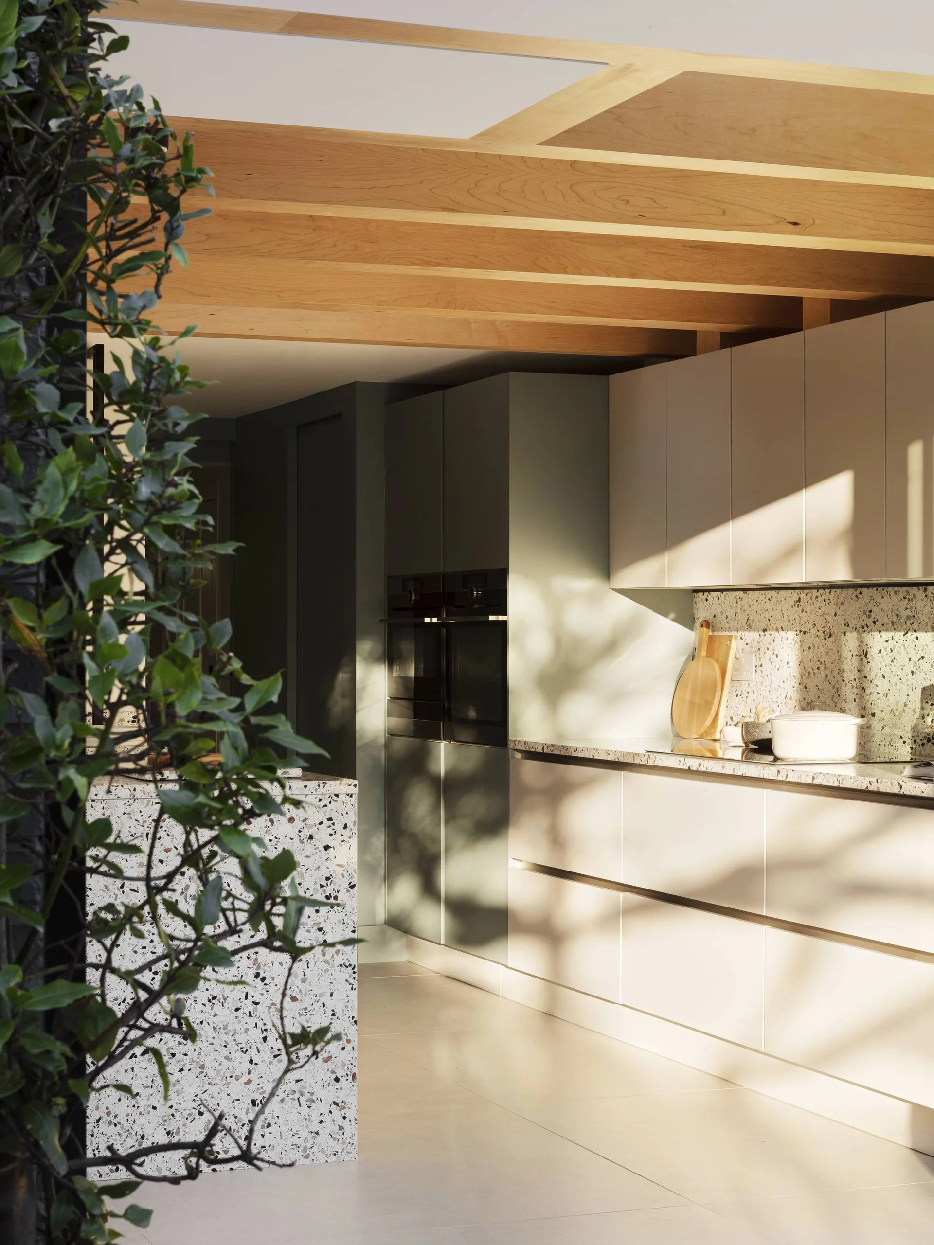 A modern kitchen features light wood ceiling beams, terrazzo countertops, and flat-panel off-white cabinets bathed in natural sunlight.