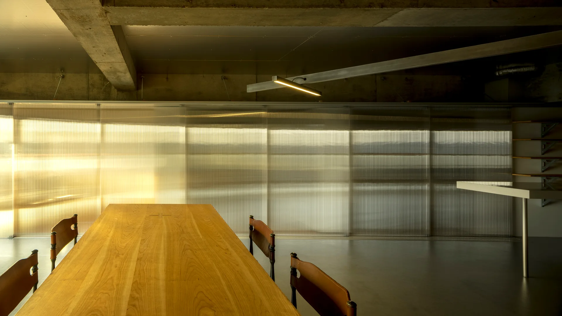 A long wooden dining table with leather-backed chairs is set against a corrugated translucent partition wall under an exposed concrete ceiling.