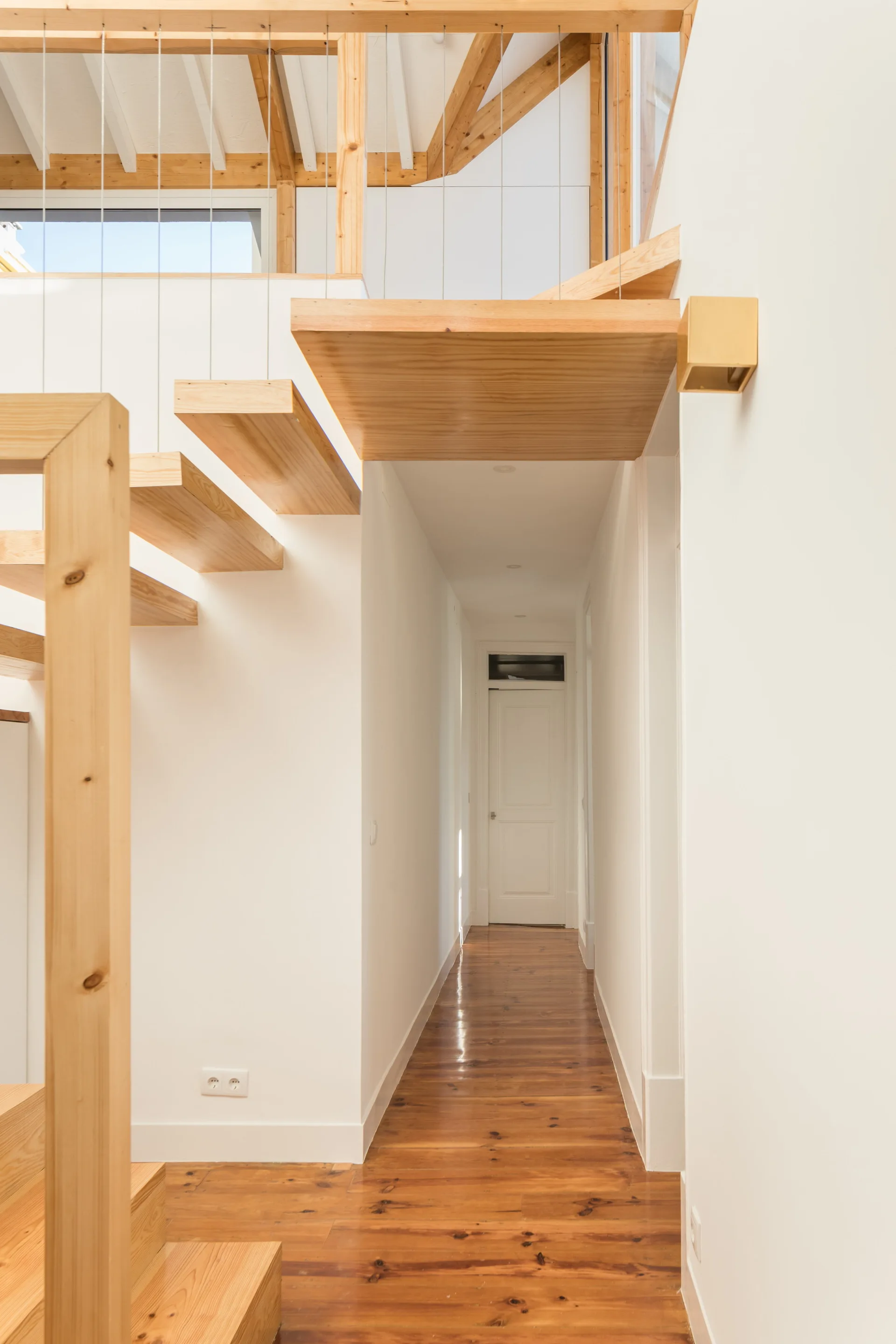 A bright white hallway featuring a floating wood staircase with a cable railing system and polished wooden floors.