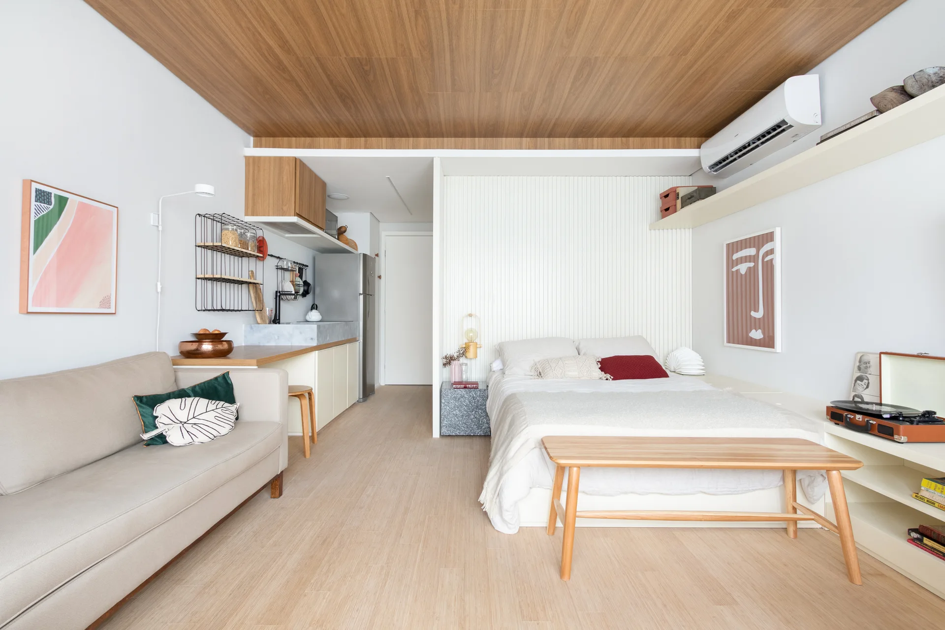 A bright studio apartment featuring a light wood paneled ceiling, a beige sofa, and a bed set against a white slatted accent wall.