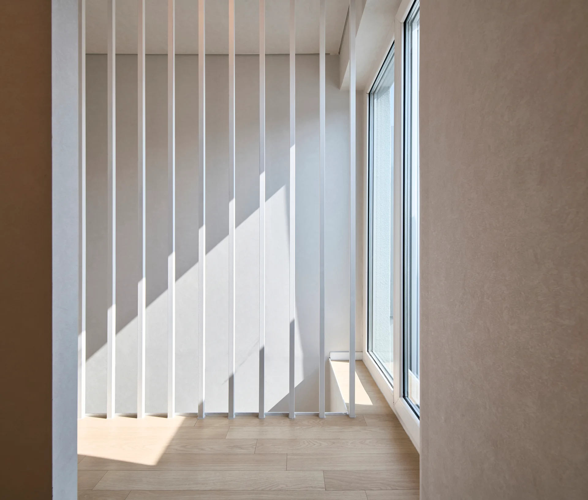 A minimalist white slatted staircase balustrade stands next to a large window on light wood floors in a sunlit interior.