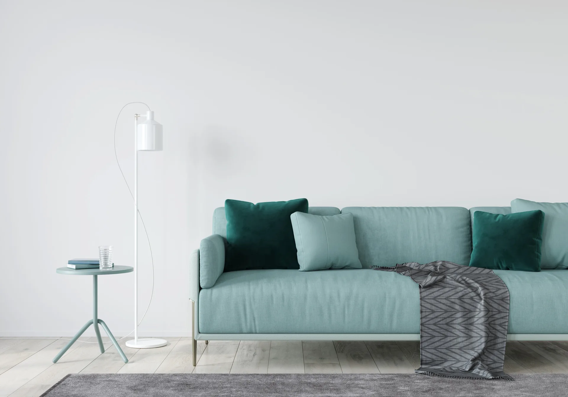 A teal sofa with dark green pillows sits in front of a white wall next to a matching round side table and white floor lamp.