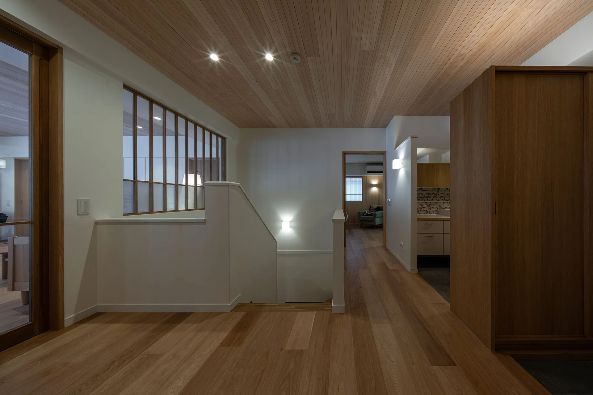 A residential landing featuring a wood plank ceiling, matching wood floors, a central staircase with an interior glass-paneled partition, and off-white walls.