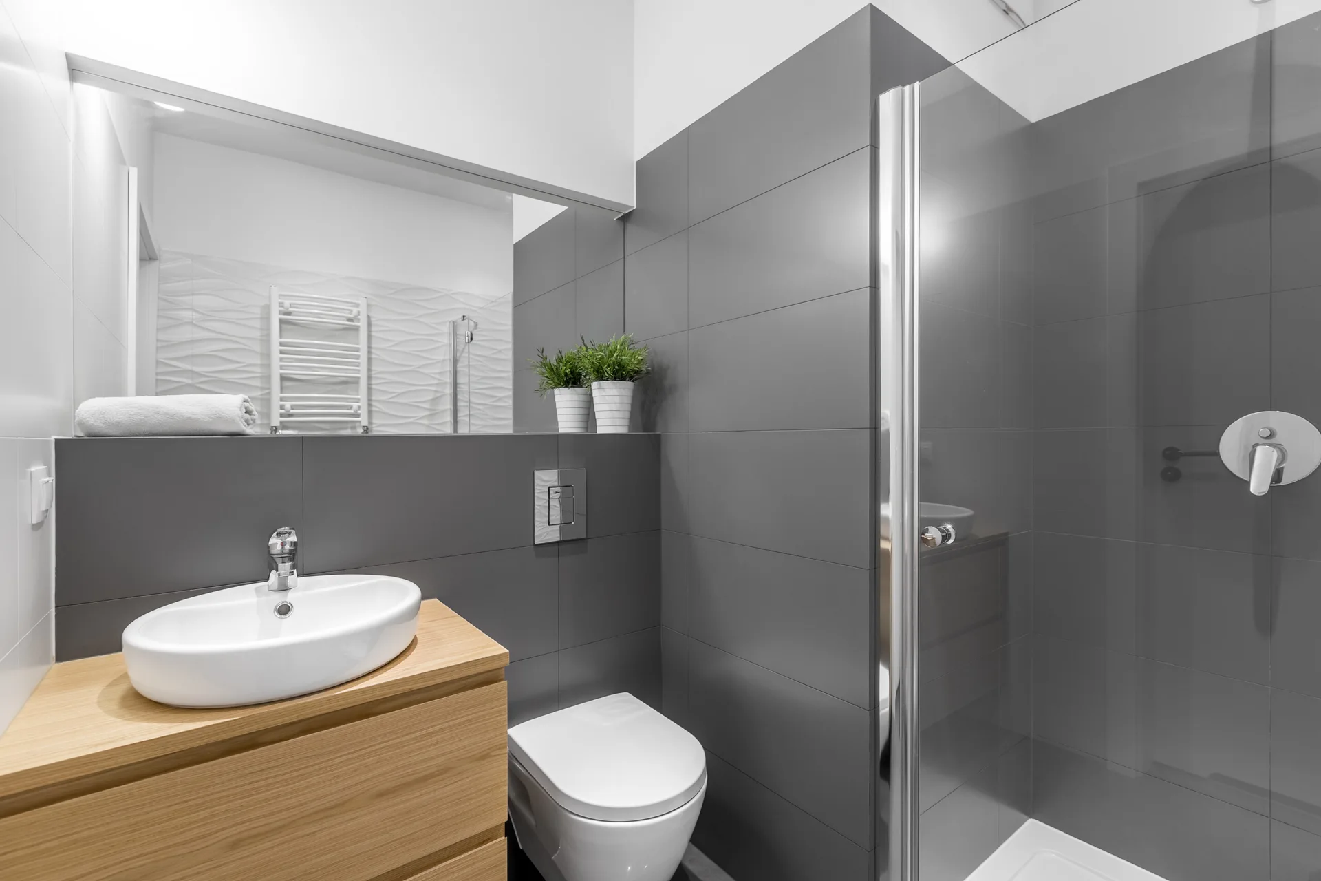 A modern bathroom featuring grey tiled walls, a wood floating vanity with a white vessel sink, a wall-mounted toilet, and a glass-enclosed shower.