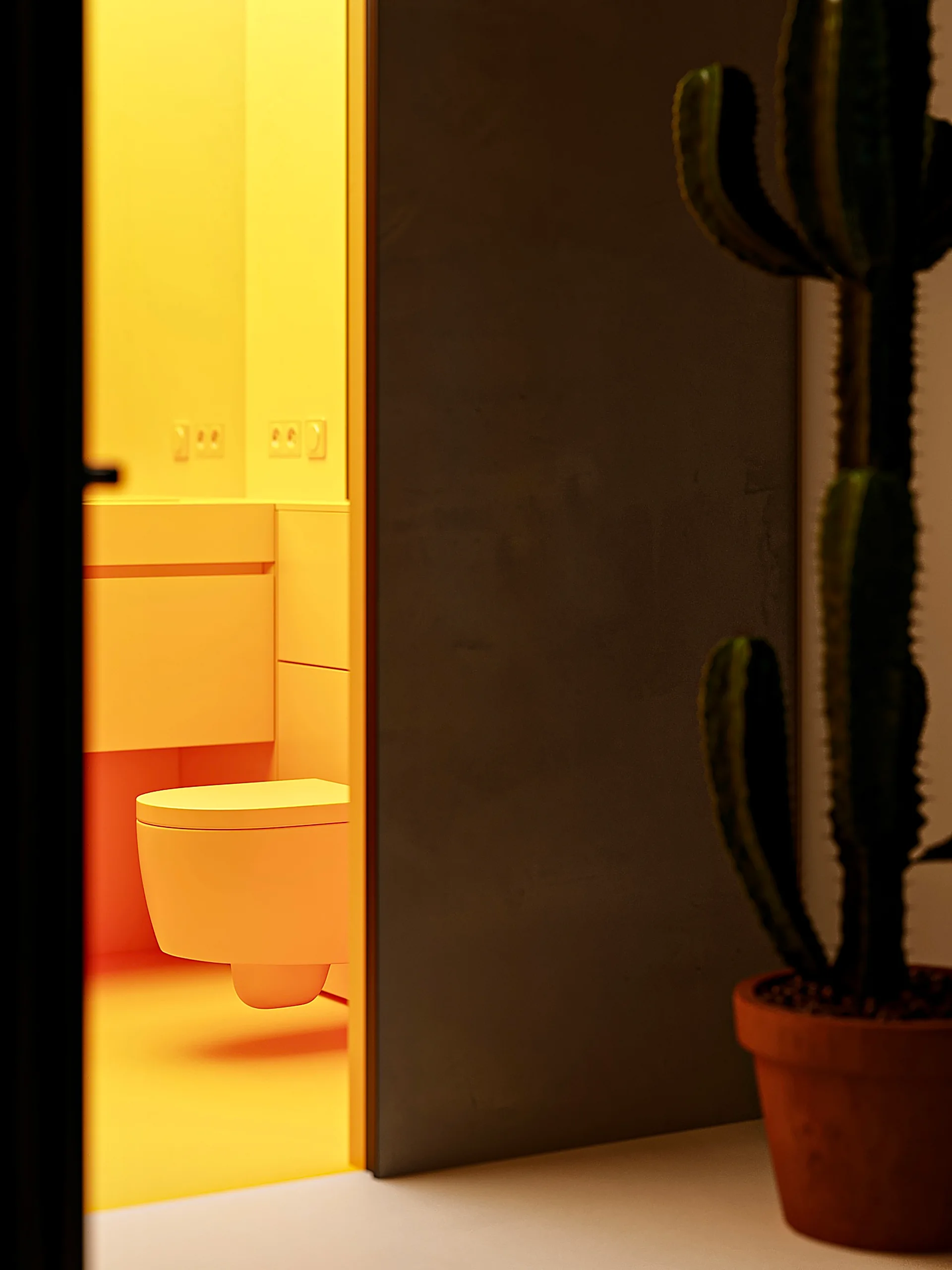 A view through a doorway into a minimalist yellow bathroom with a wall-mounted toilet, floating vanity, and a cactus in the foreground.