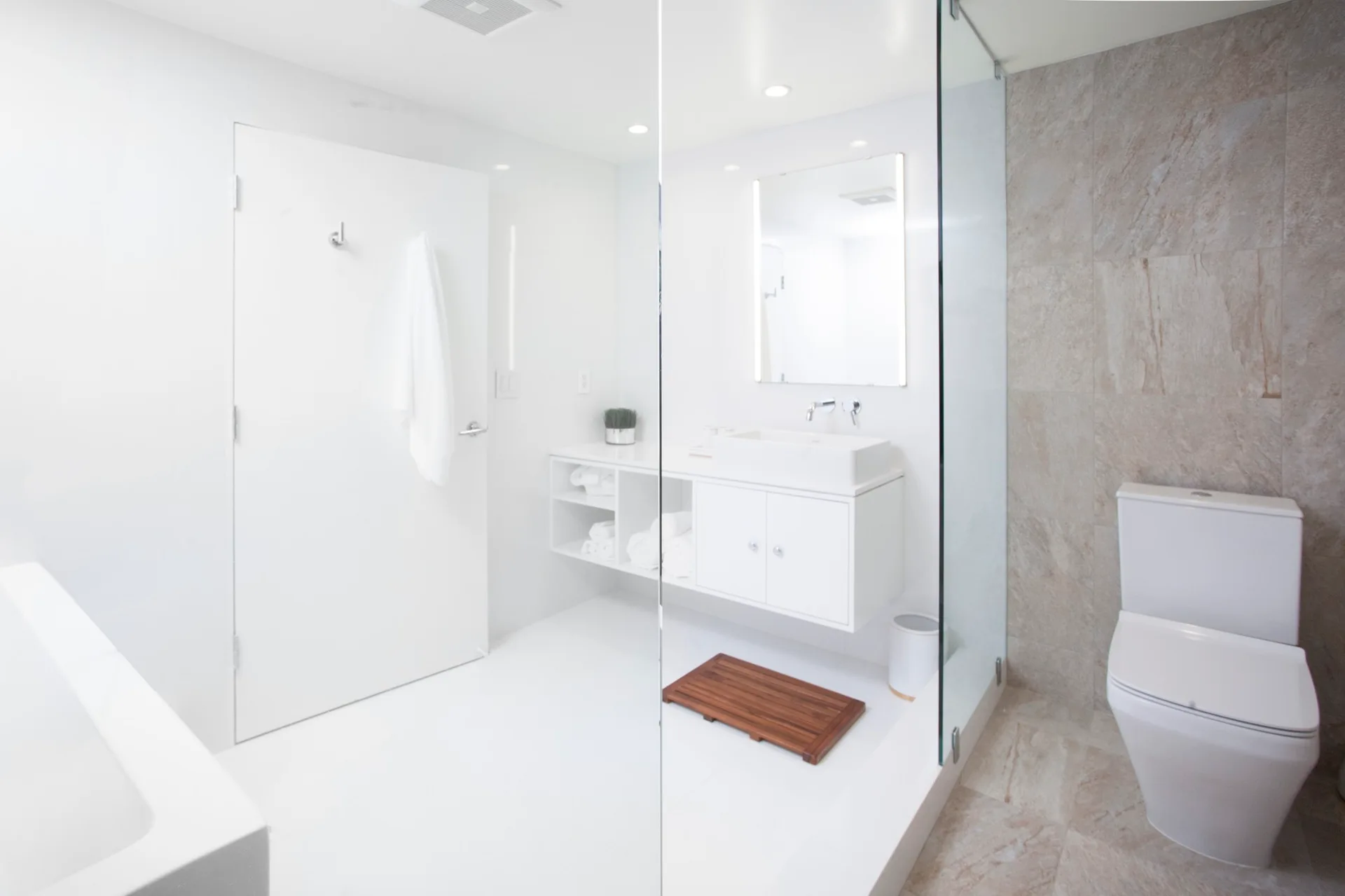 A minimalist white bathroom features a floating vanity, frameless glass shower panel, and a wooden bath mat alongside a stone-textured tiled wall.