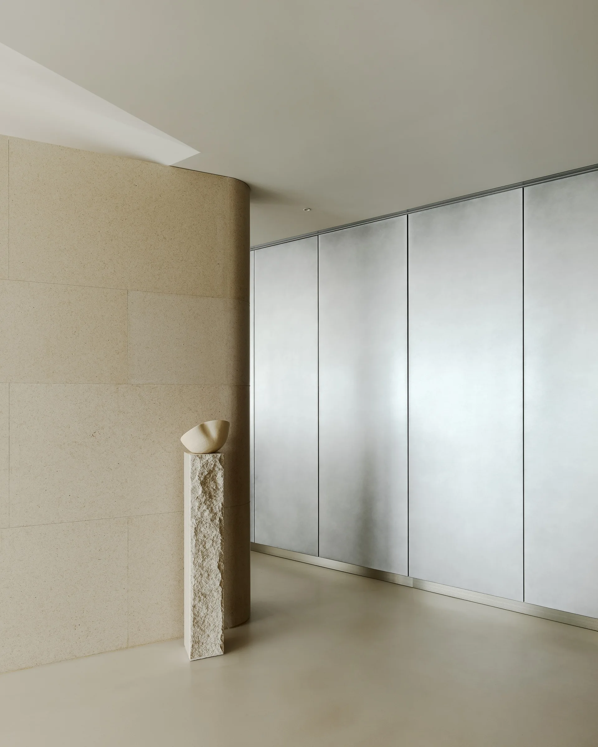 A minimalist hallway featuring a rough-hewn stone pedestal with an organic bowl, next to a curved stone-tiled wall and metallic silver cabinet doors.