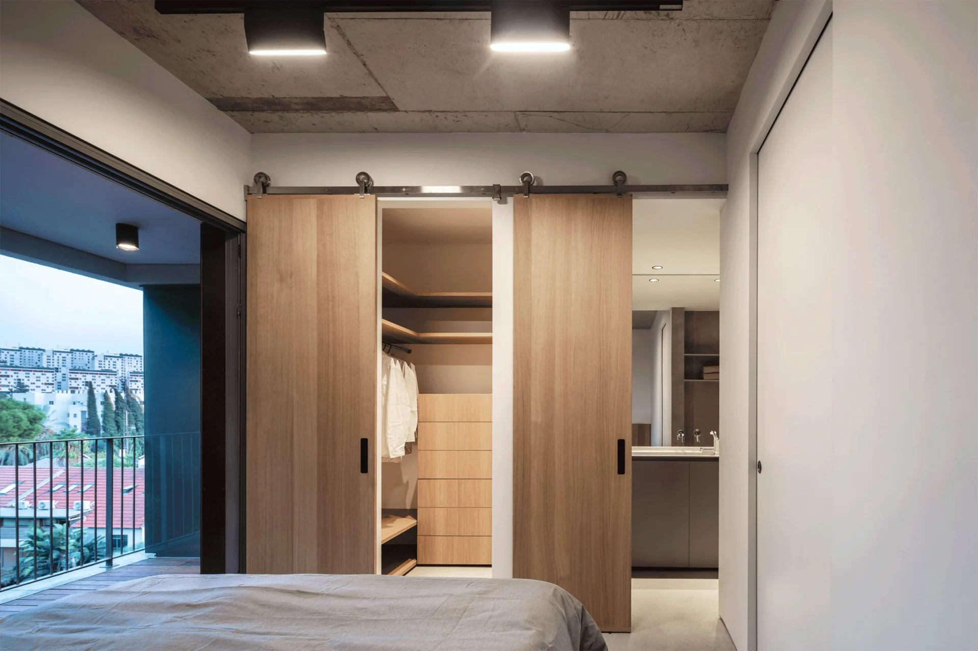 A modern bedroom featuring two sliding wooden doors on a track, an exposed concrete ceiling, and a view into a walk-in closet.