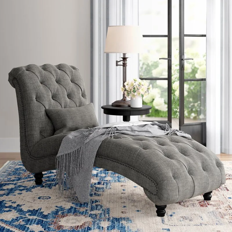 A grey tufted chaise lounge with a throw blanket stands next to a dark wood side table on a blue and white patterned rug.