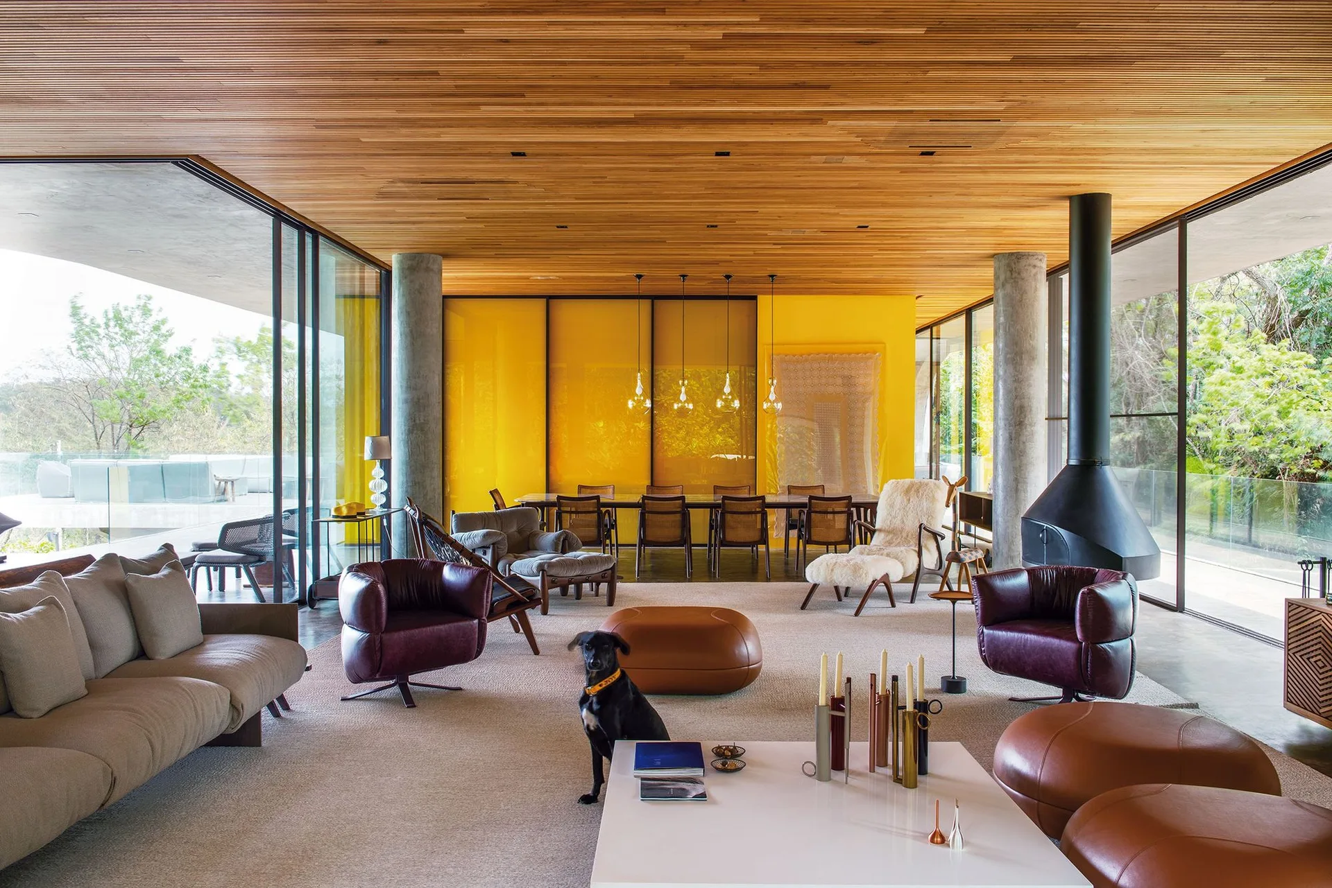 An open-concept living and dining space features a wood slat ceiling, a bright yellow glass partition, concrete pillars, and mid-century modern seating.