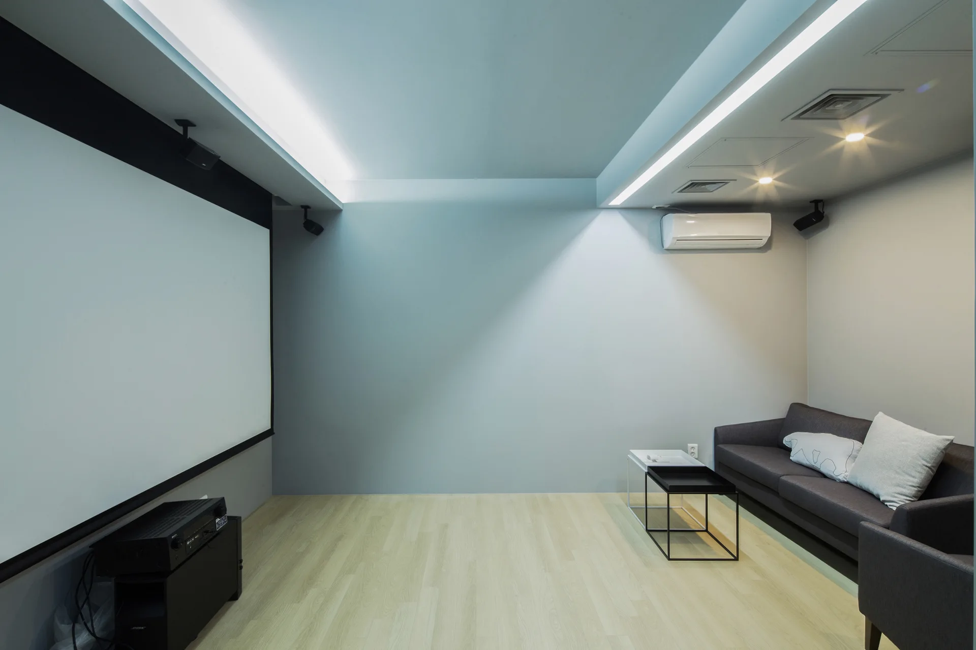 A minimalist media room features a large white projection screen, a grey sofa, and light wood flooring under recessed linear ceiling lights.