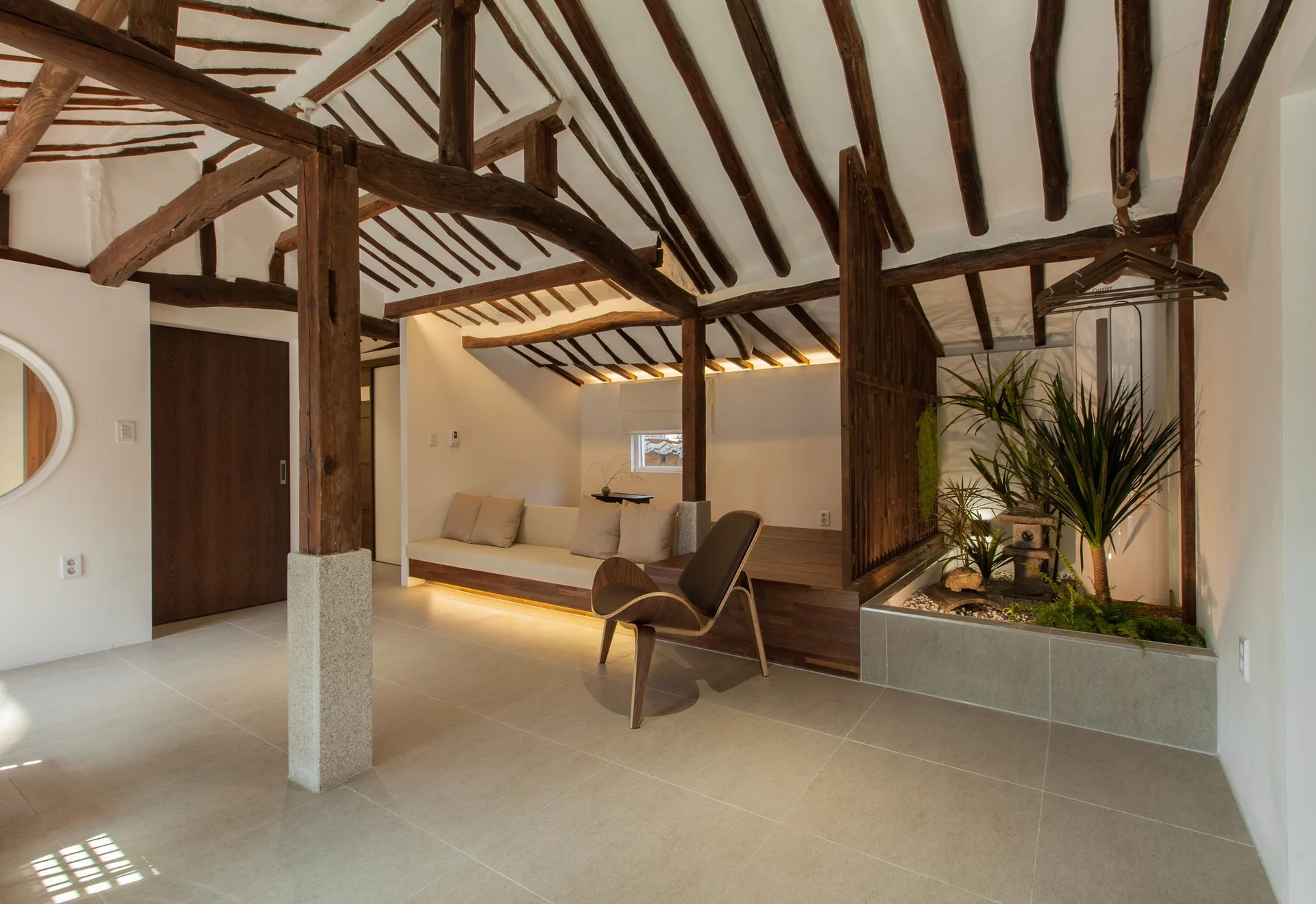 A minimalist living room featuring a traditional exposed dark wood rafter ceiling, built-in low bench seating, a curved lounge chair, and an indoor planter.