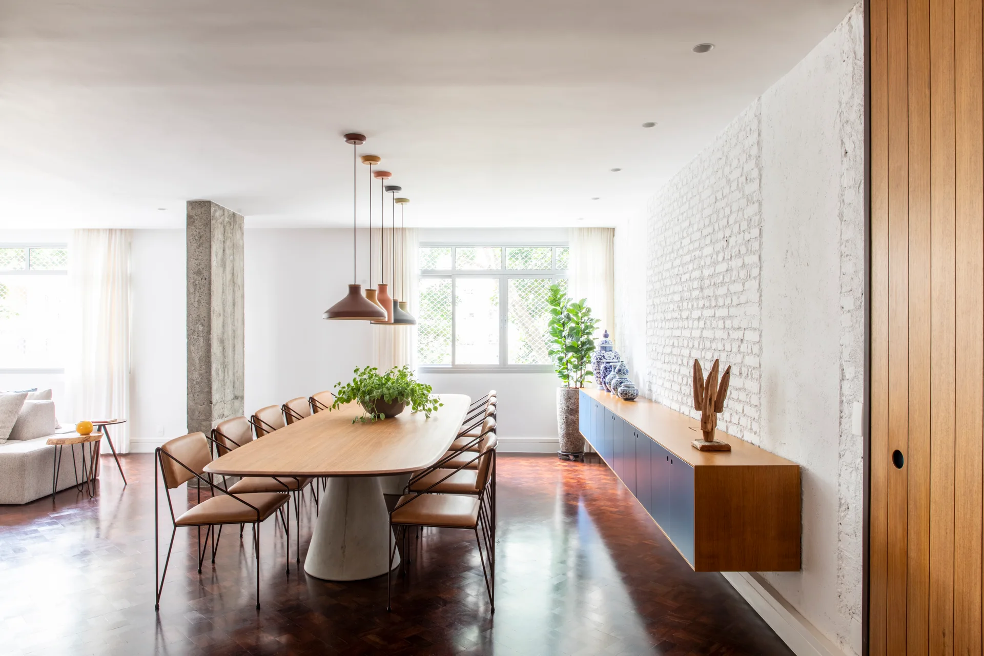 A spacious dining room features a long wooden table with leather chairs, a white exposed brick wall, and a long floating blue sideboard.