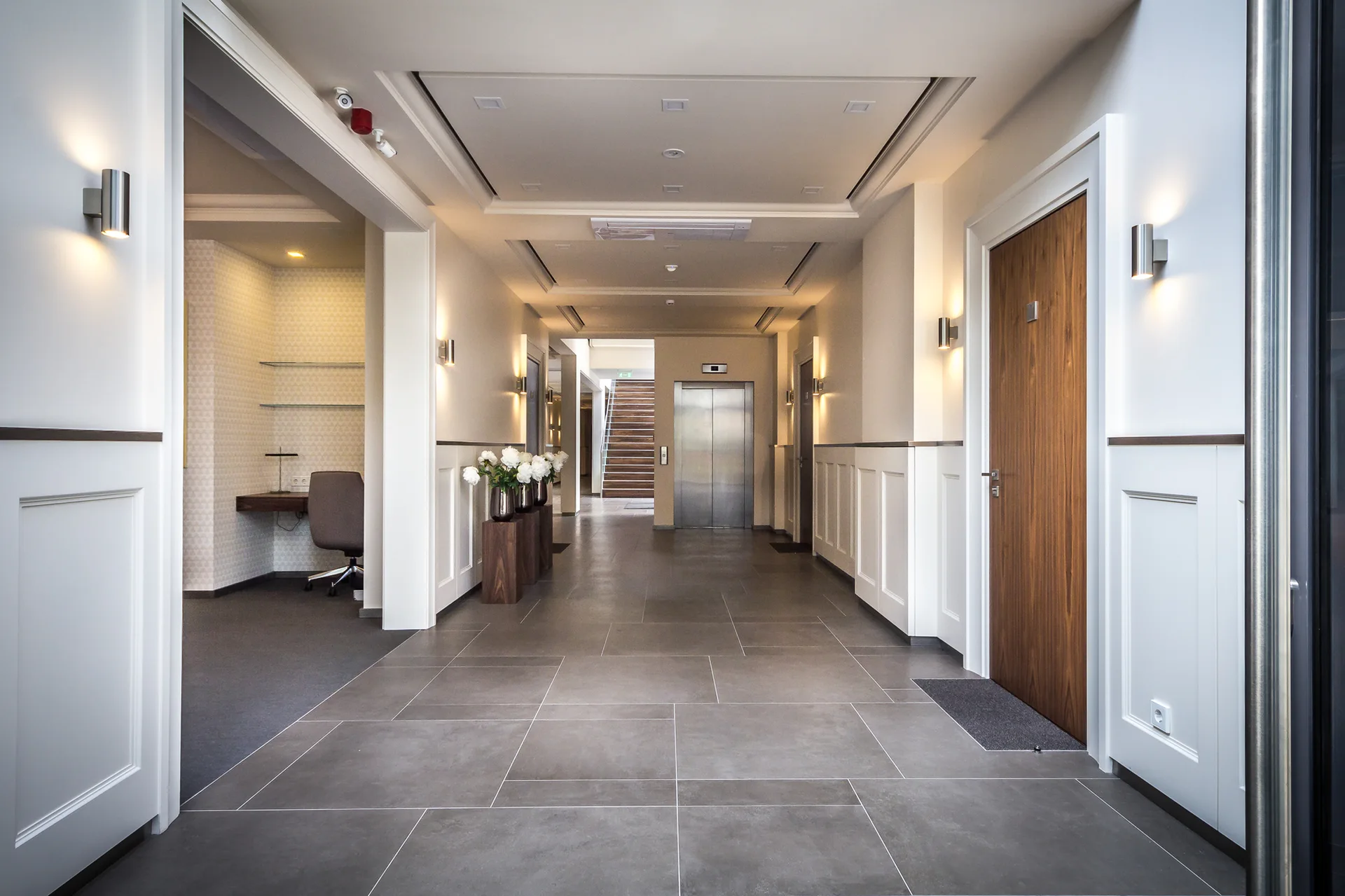 A modern building hallway features gray tile flooring, lower white wainscoting, cylinder wall sconces, and a wood door near an elevator.