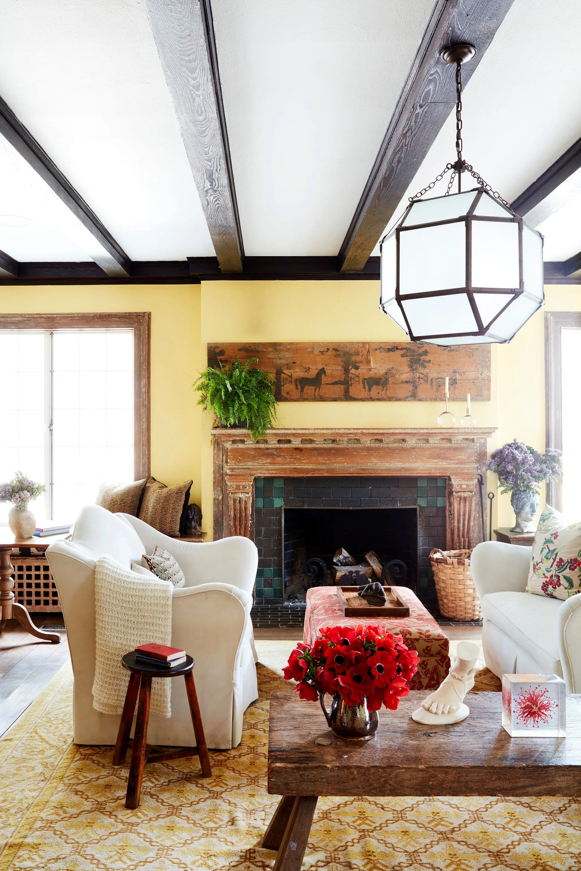 A bright yellow living room features white armchairs, a rustic wood fireplace mantel, a low wooden coffee table, and dark exposed ceiling beams.