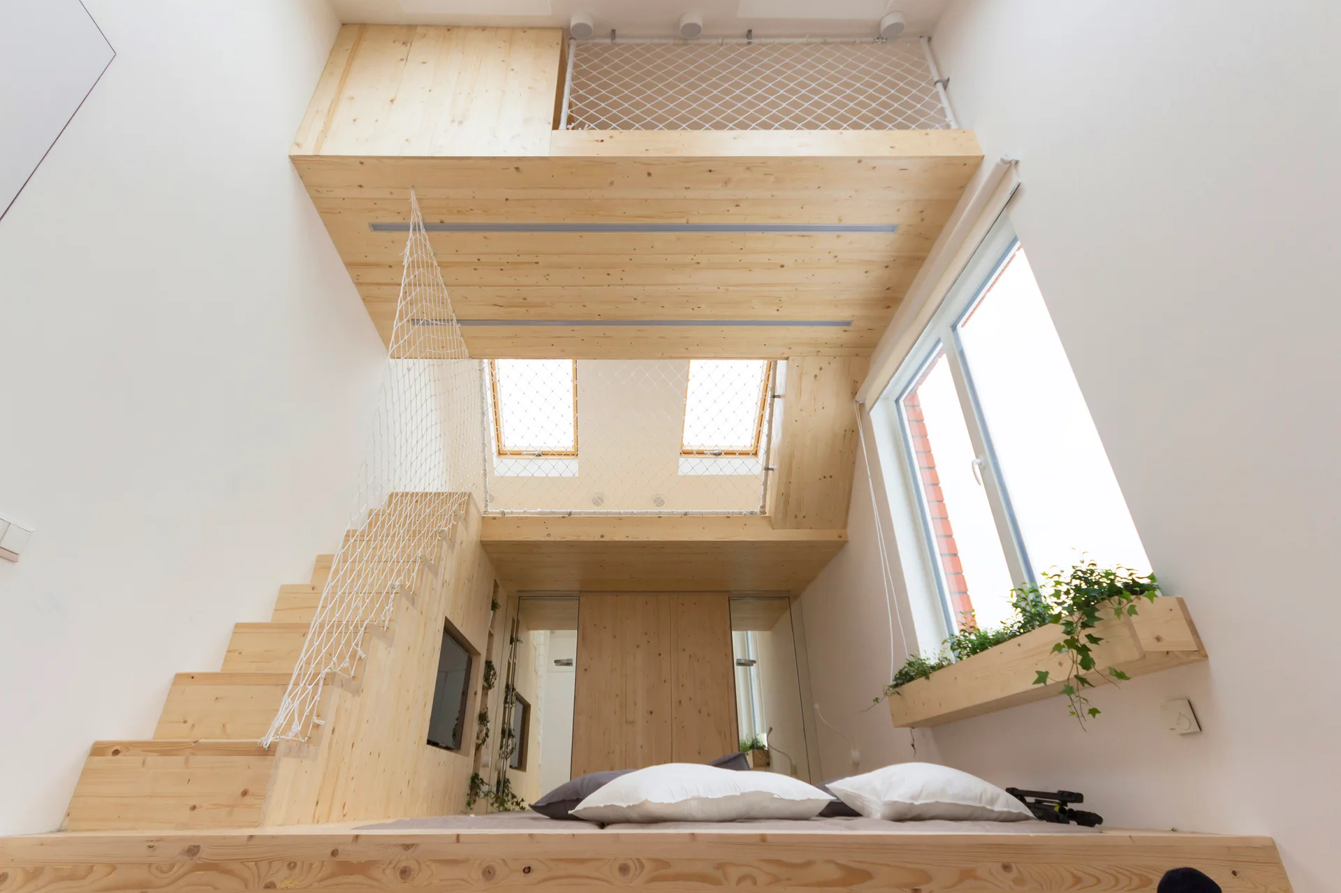 A compact bedroom built from light wood features a platform bed, stairs with white safety netting, a mezzanine loft, and overhead skylights.