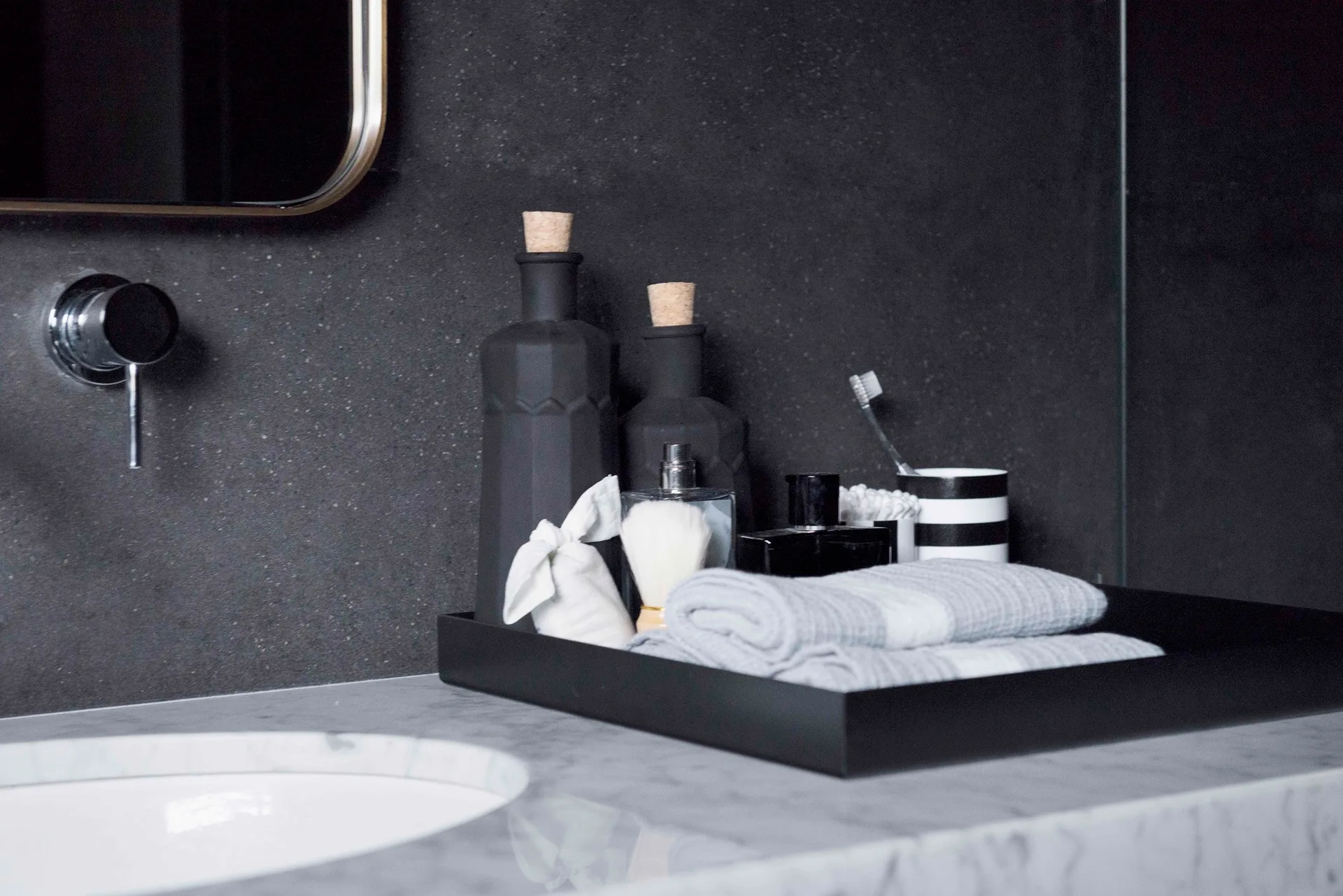 A close-up of a marble bathroom vanity featuring a wall-mounted faucet and a black tray holding grooming accessories and towels against a dark wall.
