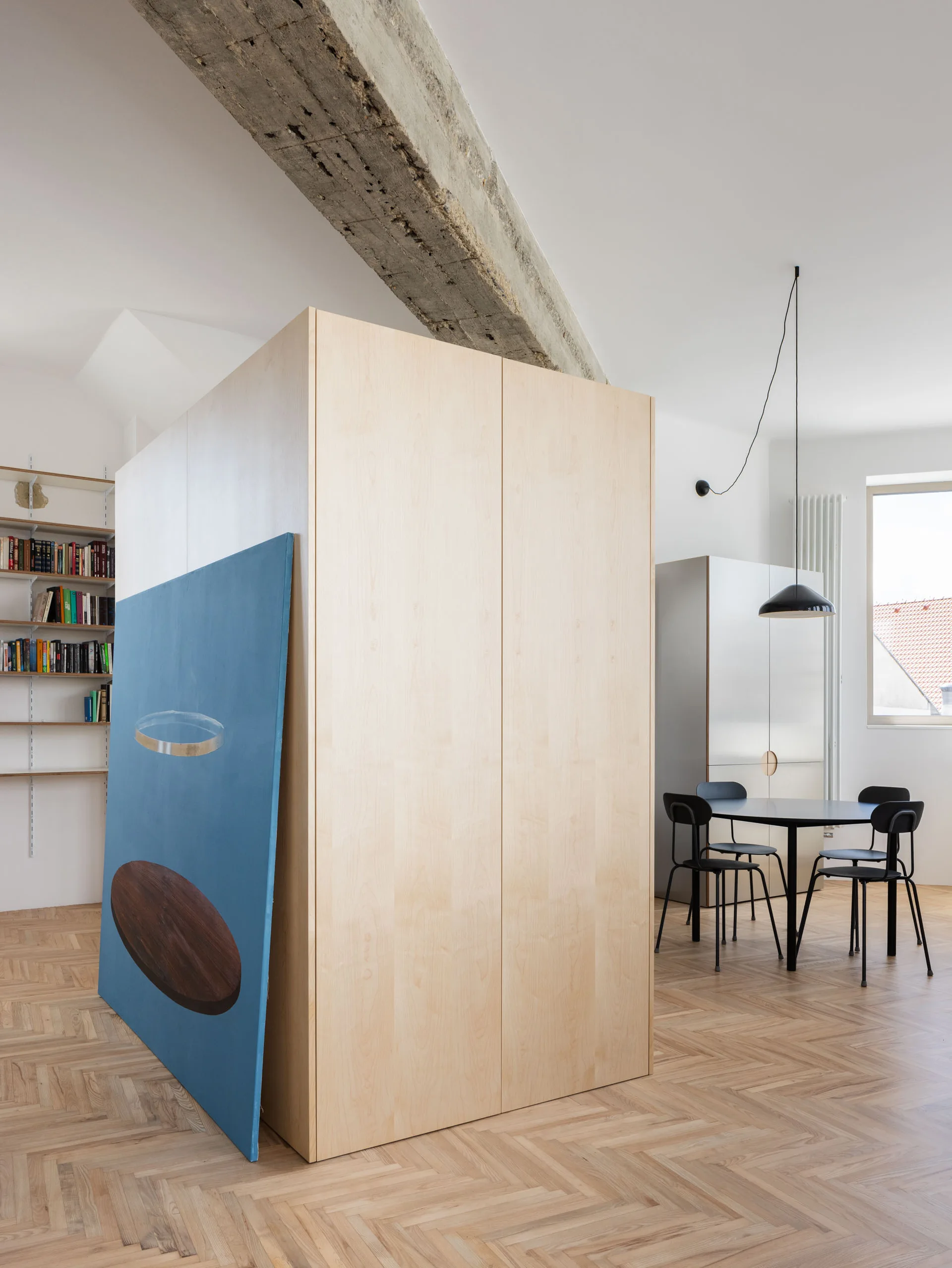 A large light wood room divider stands on herringbone flooring in a minimalist space with an exposed concrete beam.