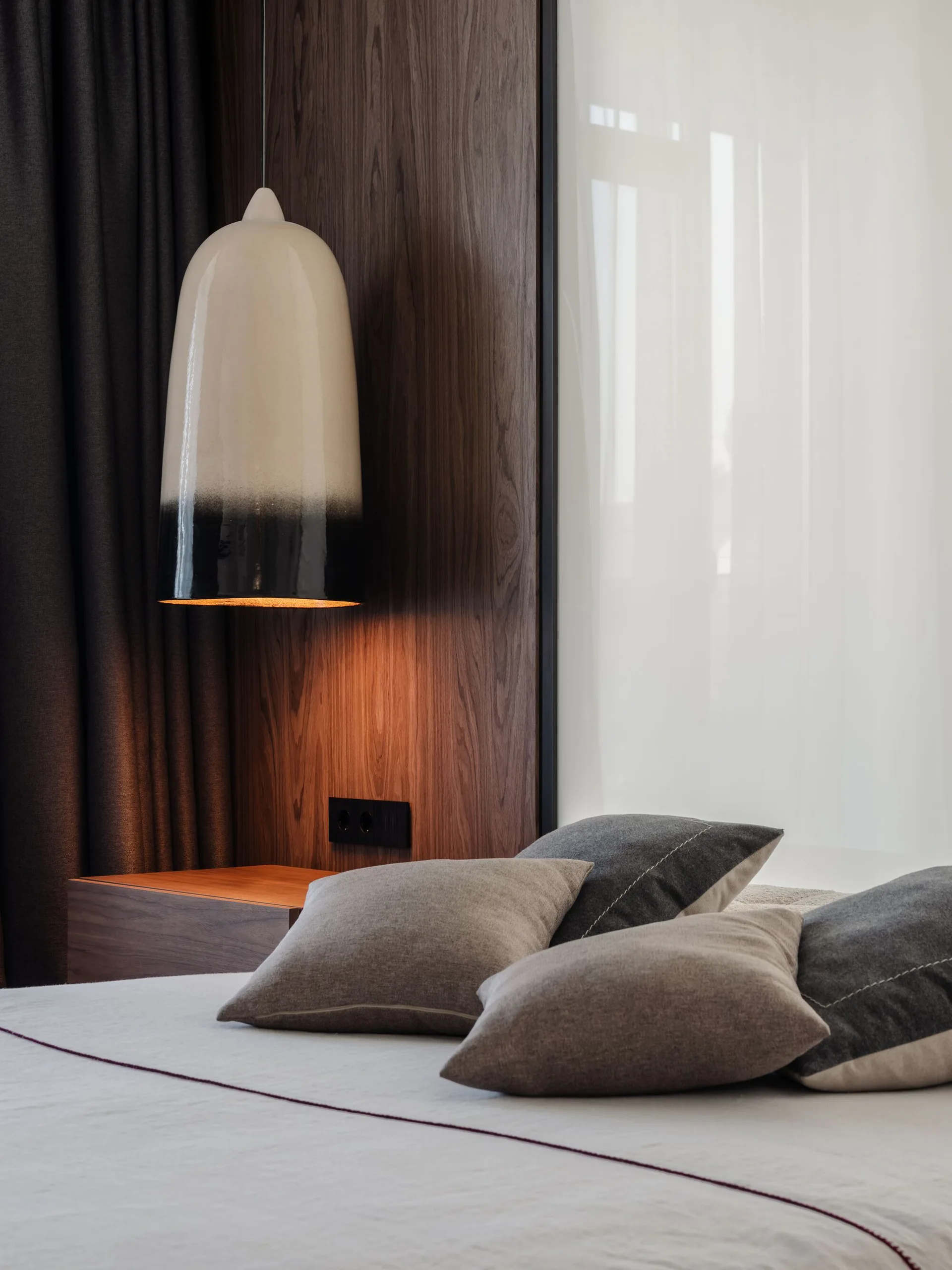 A modern bedroom corner featuring a two-toned ceramic pendant light hanging next to dark wood wall paneling and a bed with grey pillows.