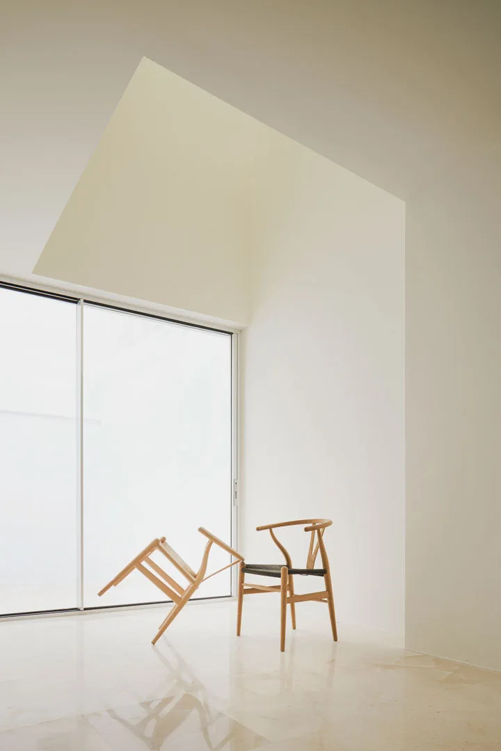 Two light wood wishbone chairs sit on a polished stone floor next to a large sliding glass door in a minimalist room with white walls.