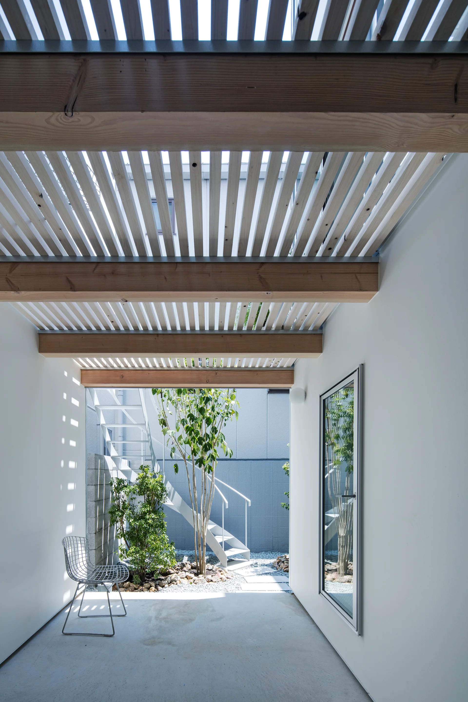 A minimalist outdoor patio features a slatted wooden canopy overhead casting striped shadows onto a white wall next to a small gravel courtyard garden.