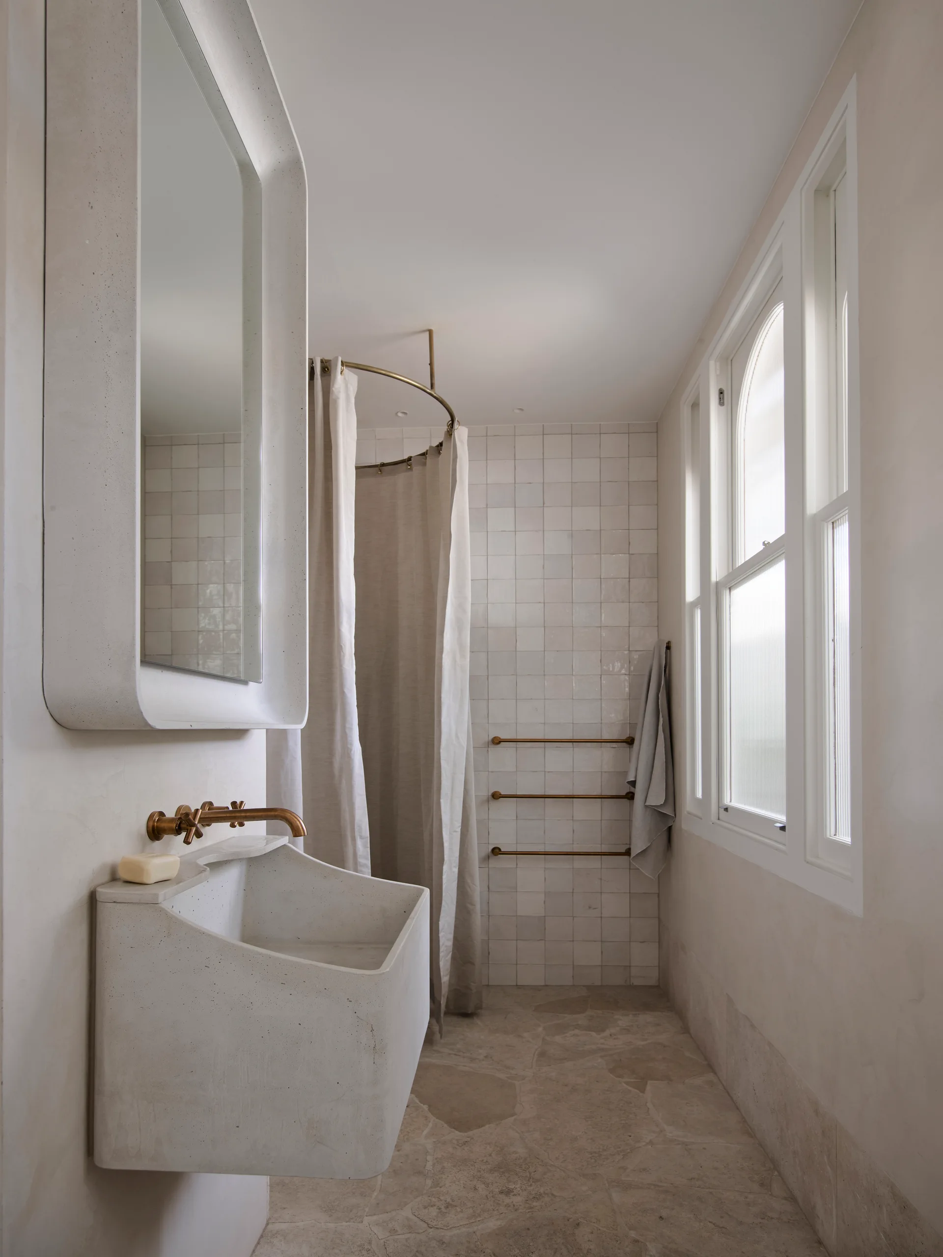 A neutral plaster bathroom features a wall-mounted stone sink, a rounded mirror, a brass faucet, and an arched window.