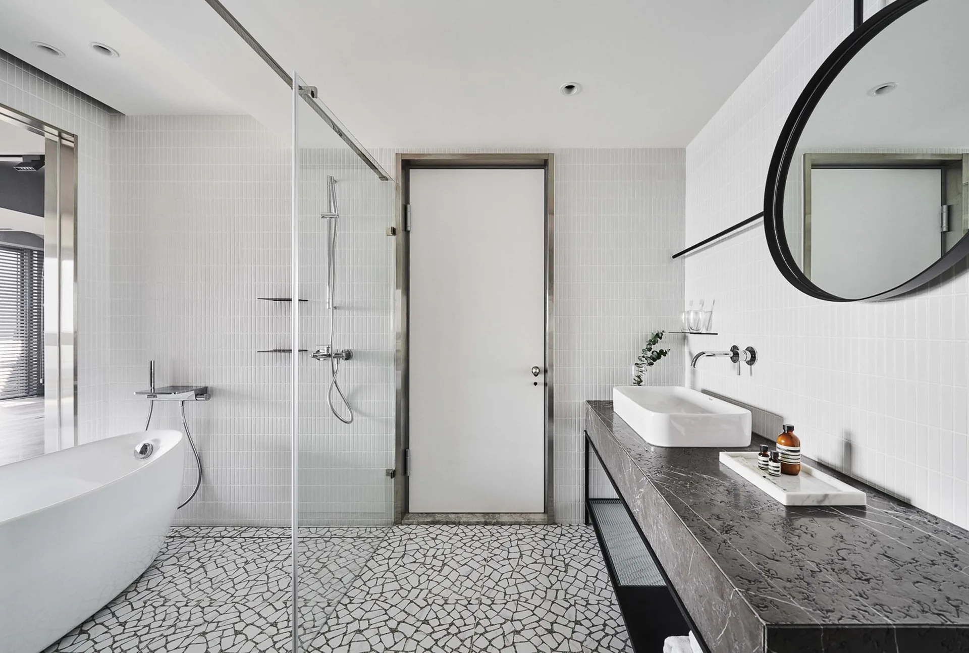 A modern white bathroom features a stone mosaic tile floor, a dark floating vanity with a round mirror, a glass-walled shower, and a freestanding tub.