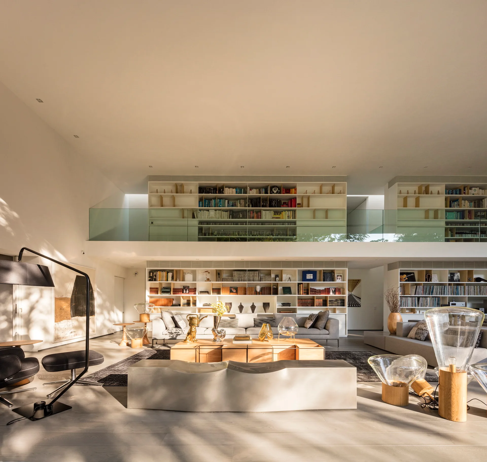 A double-height living room features grey sofas, a modular wooden coffee table, and an upper mezzanine library with glass railings and white bookshelves.