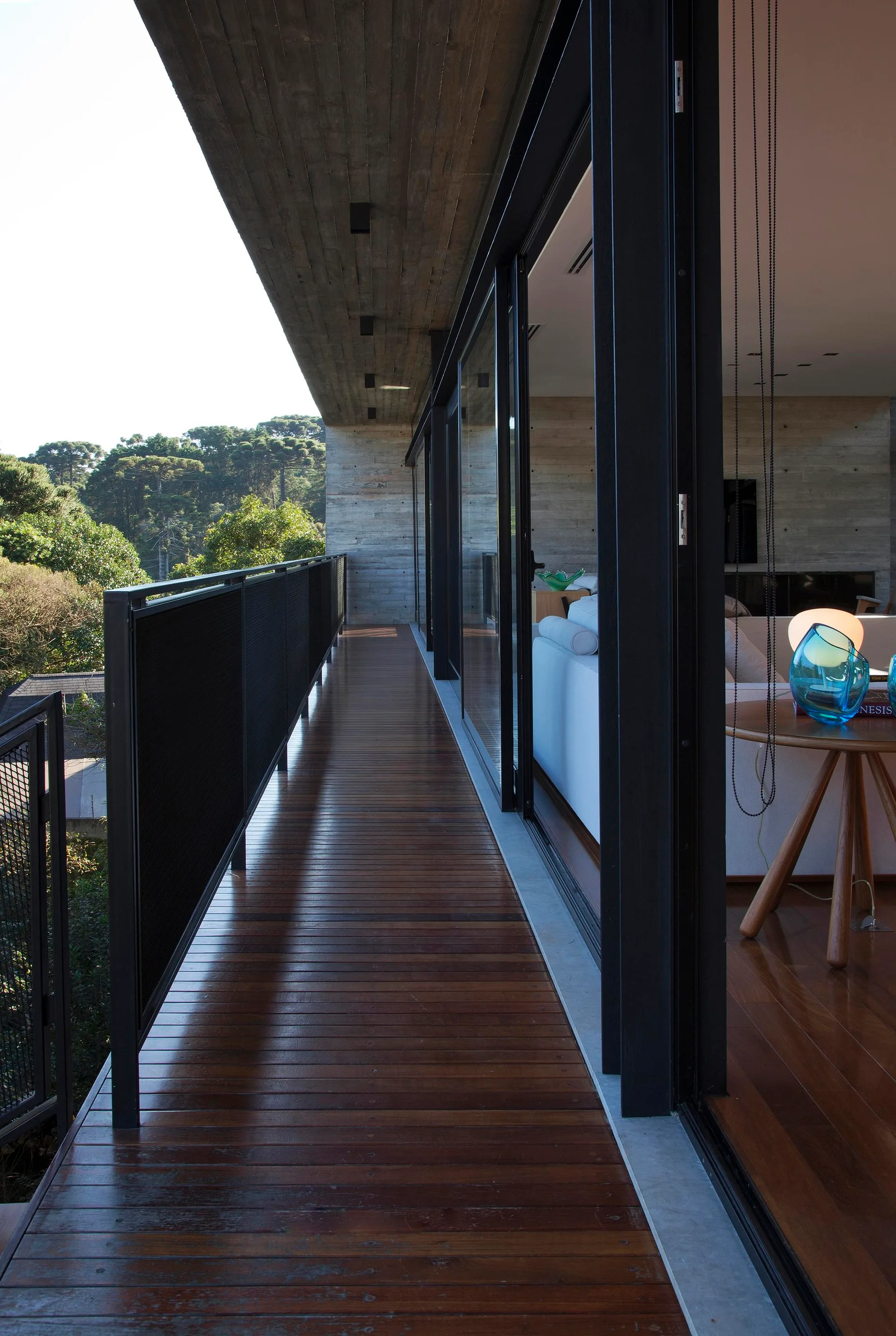 A long wood deck balcony with a black metal railing features an exposed concrete ceiling and glass sliding doors looking inside.