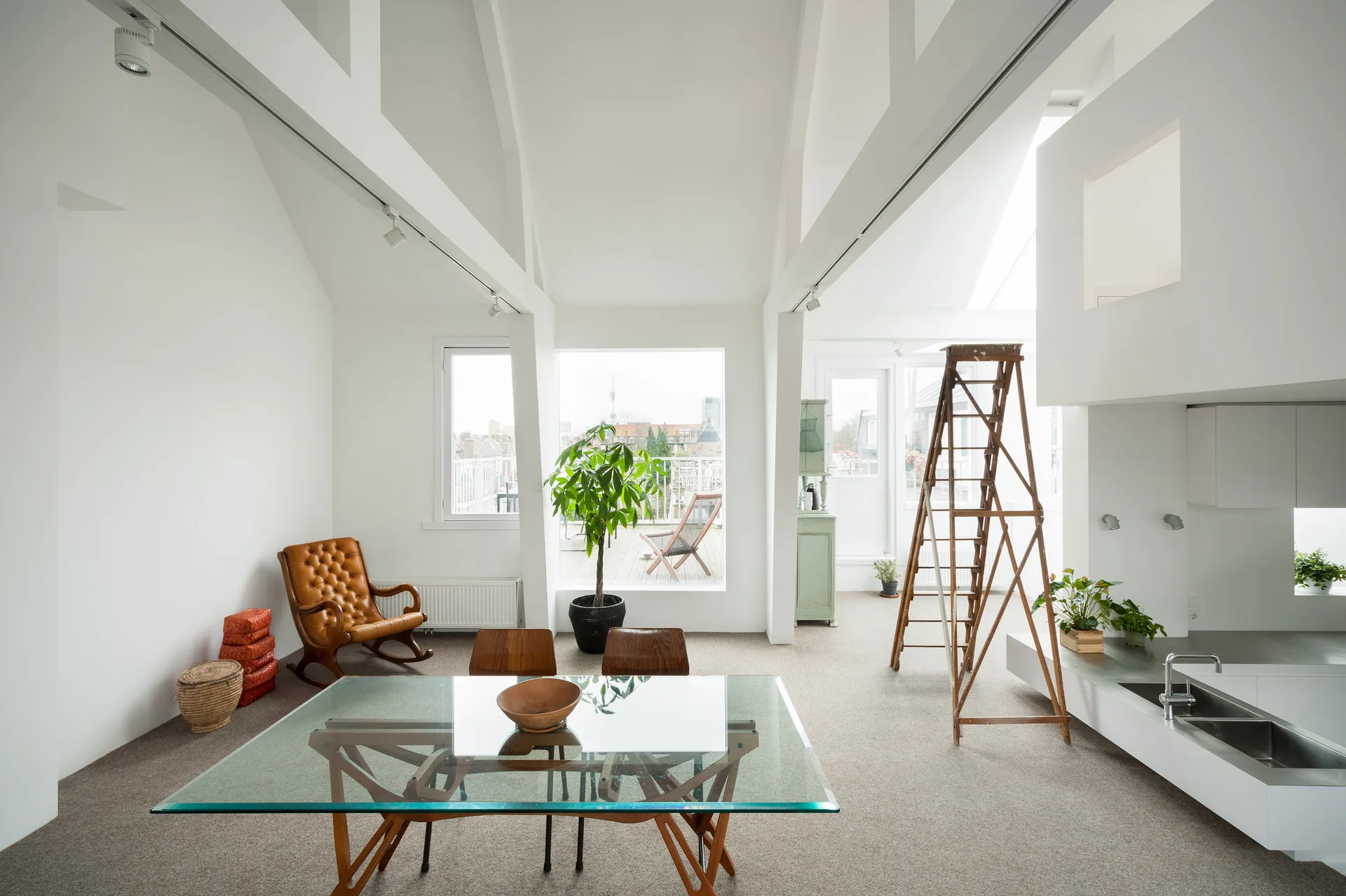A bright loft living space with white vaulted ceilings, a glass dining table, a leather rocking chair, and a decorative wooden ladder.