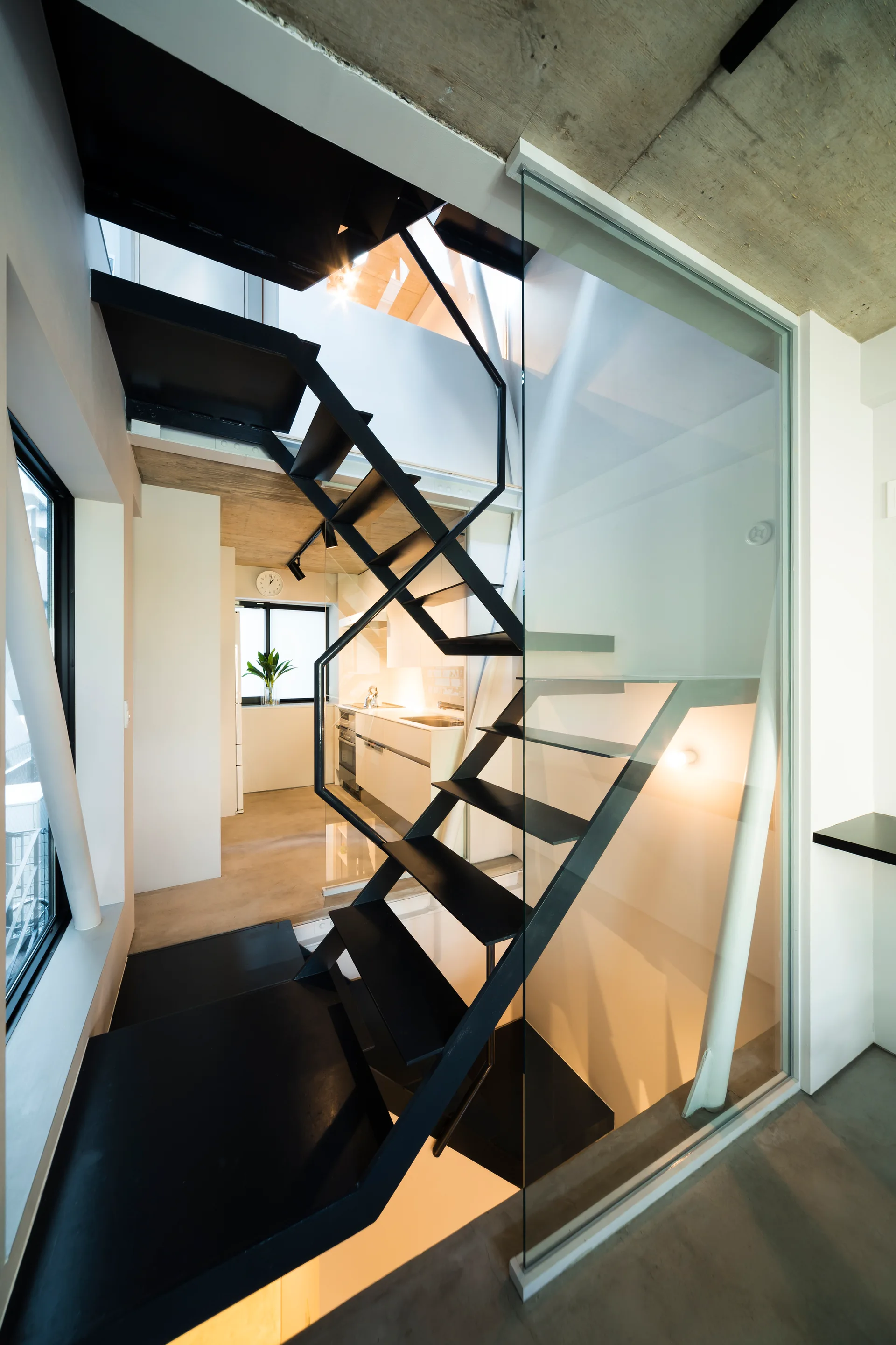 An industrial black metal staircase with open treads is enclosed by a glass partition next to an exposed concrete ceiling and a white kitchen.