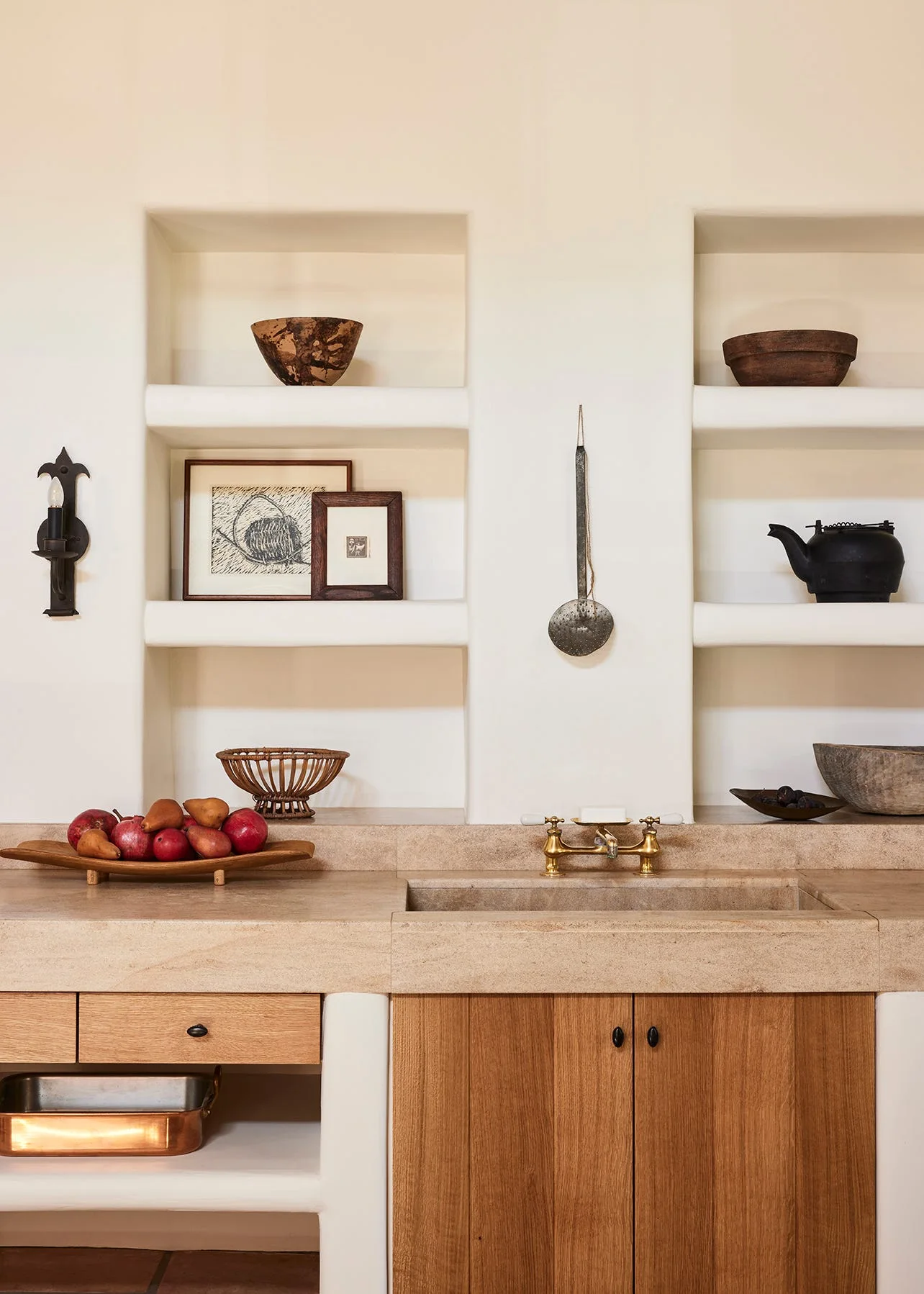 A rustic kitchen features plaster wall niches with decorative objects above a honed stone countertop, integrated sink, brass faucet, and wood cabinets.