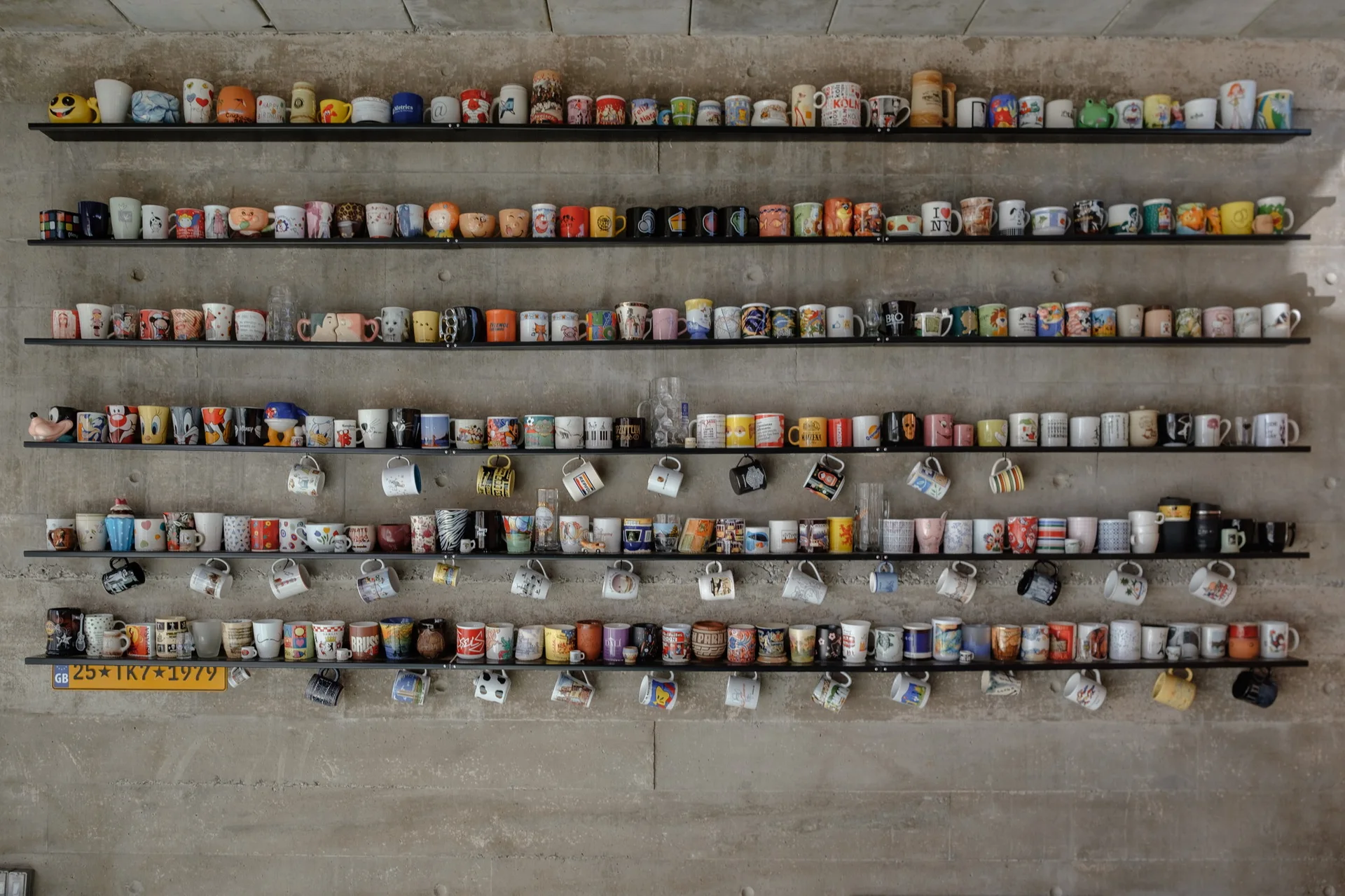 Multiple black metal shelves mounted on a raw concrete wall display a large, colorful collection of ceramic coffee mugs.