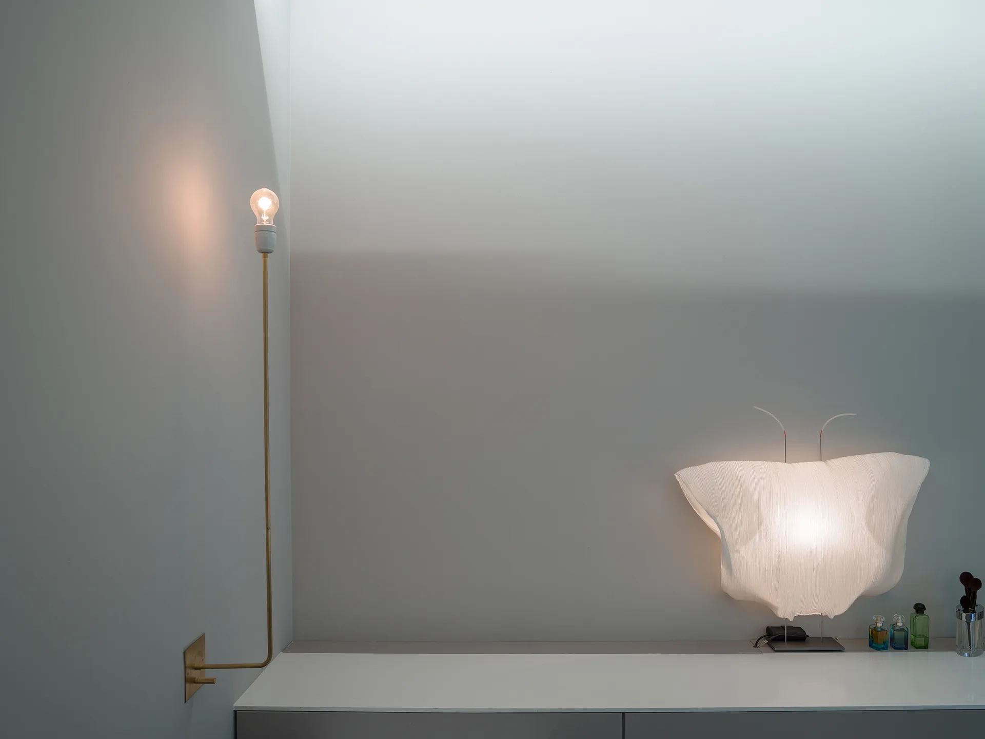 A thin brass wall sconce with a bare bulb and a sculptural pleated table lamp sit on a white console table against a grey wall.