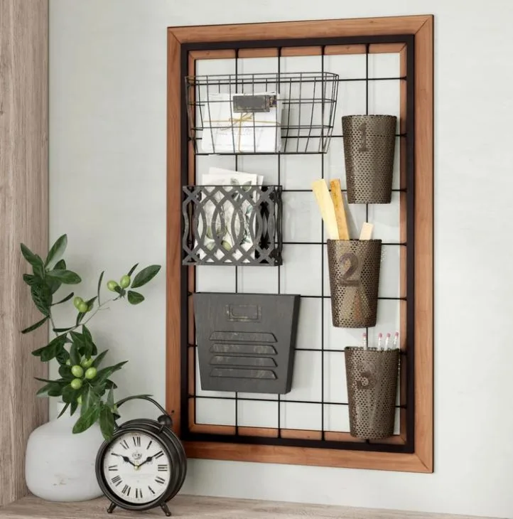 A rectangular wood-framed black wire grid organizer with hanging metal storage bins mounted on a wall next to a clock and plant.