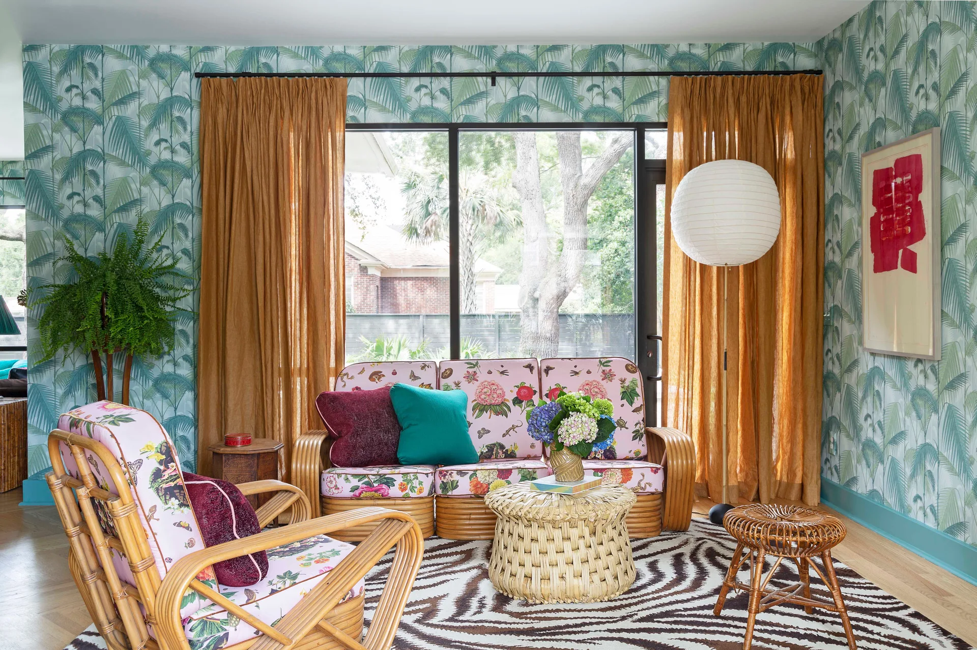 A tropical living room features a rattan sofa with floral cushions, green palm leaf wallpaper, orange curtains, and a zebra print rug.