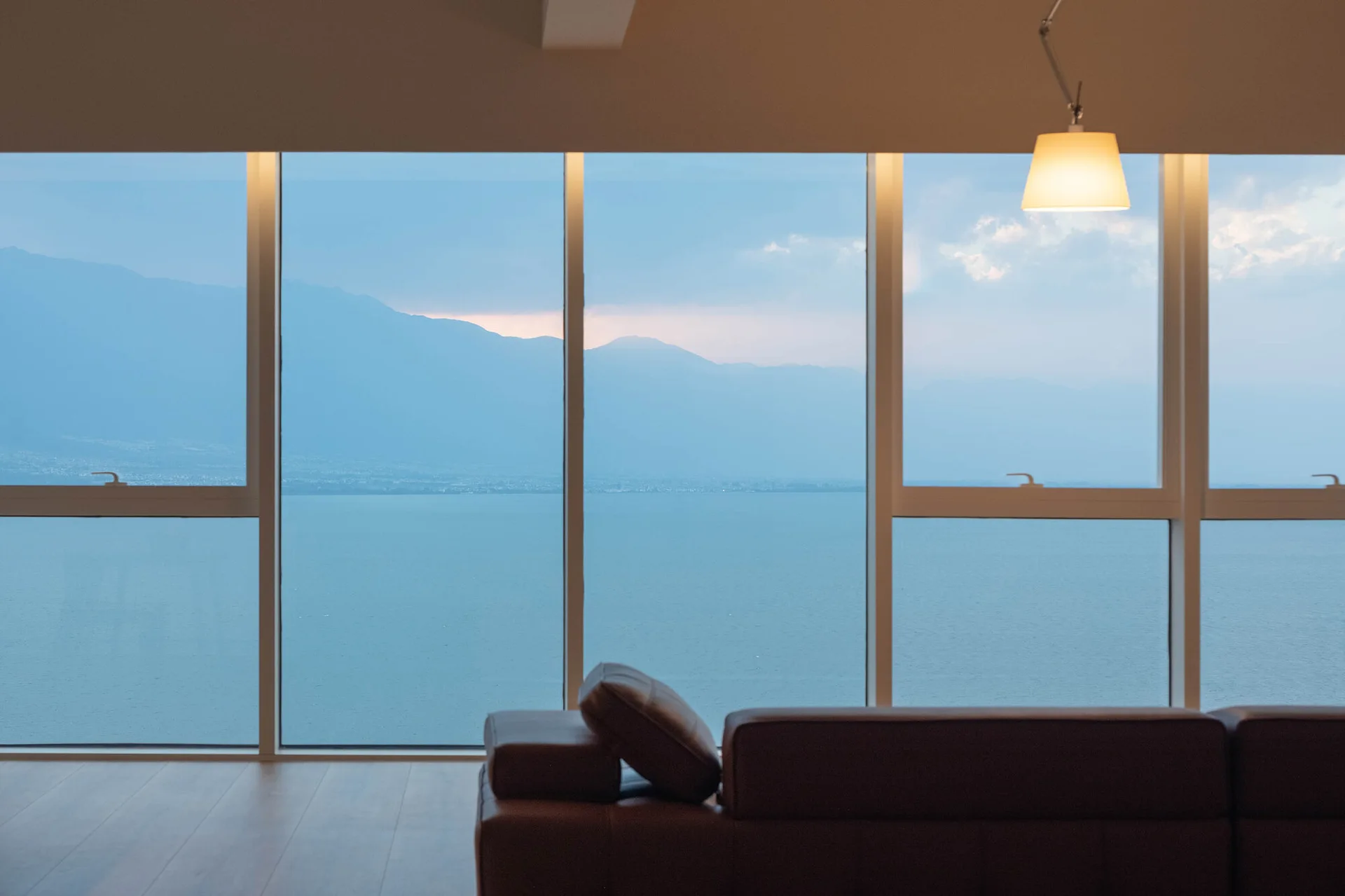 A low brown leather sofa sits in a minimalist room in front of large windows overlooking a peaceful blue lake and distant mountains at dusk.